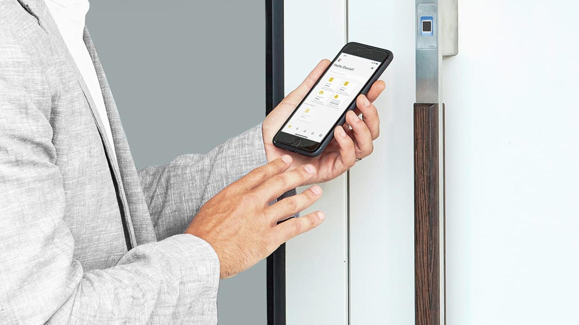 Eine Person in einem hellgrauen Anzug benutzt eine Smartphone-App, um ein modernes digitales Türschloss mit einem Fingerabdruckscanner an einer weißen Tür zu entriegeln.