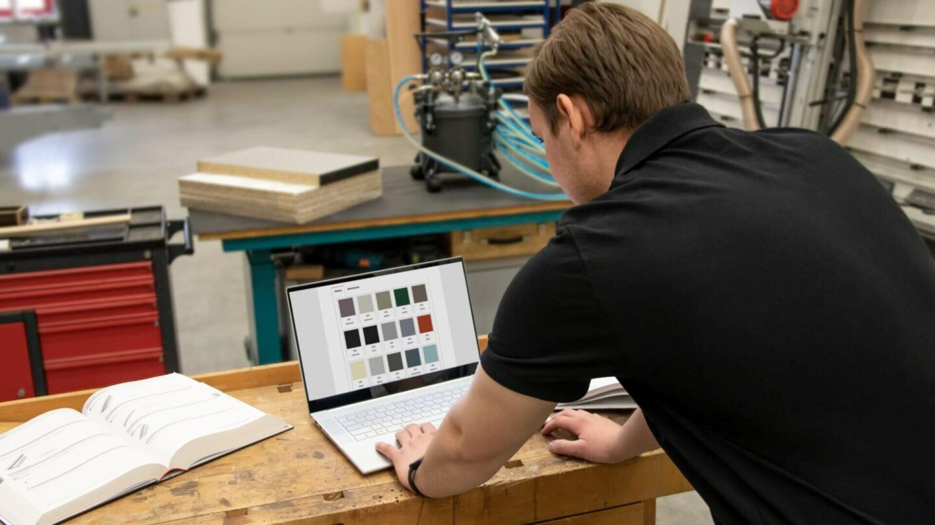 Eine Person in einem schwarzen Hemd steht an einer Werkbank und schaut auf einen Laptop, der Farbmuster anzeigt. Auf dem Tisch liegen aufgeschlagene Bücher und Materialien, und im Hintergrund ist eine Werkstattausrüstung zu sehen.