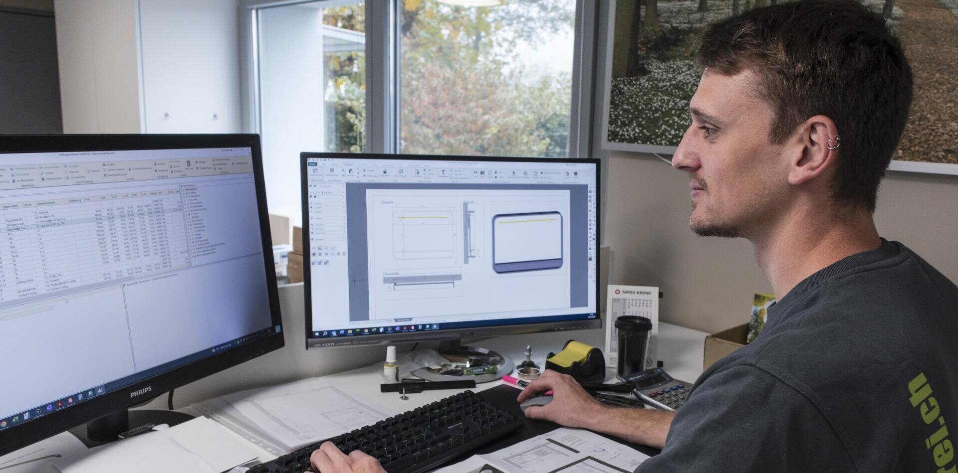 Un homme est assis à un bureau et travaille sur un ordinateur doté de deux écrans, l'un affichant une feuille de calcul et l'autre un dessin technique. Des papiers, un clavier et des fournitures de bureau sont posés sur le bureau. Une fenêtre montre des arbres à l'extérieur.