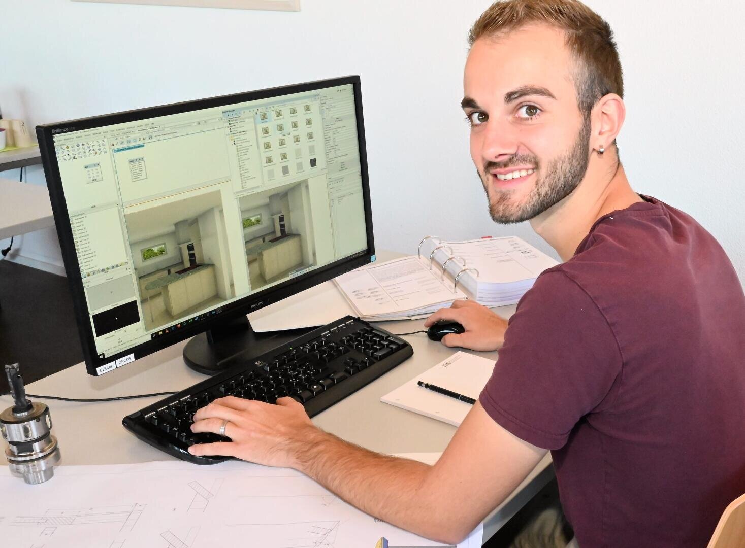 Eine Person mit kurzen Haaren und Bart, die ein kastanienbraunes Hemd trägt, sitzt an einem Schreibtisch mit einem Computer und einer Software für architektonische Entwürfe, umgeben von Papieren, einer Mappe und einem Notizblock, und lächelt in die Kamera.