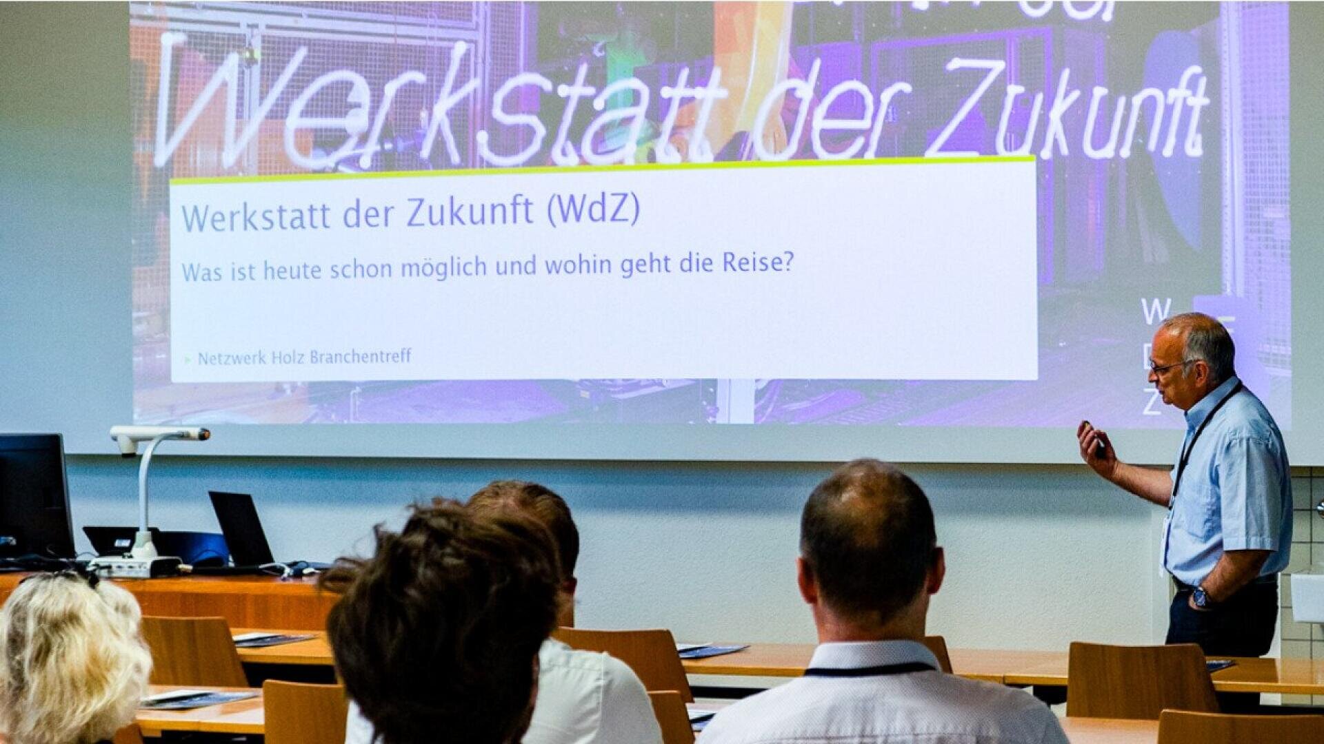 Un homme se tient devant une salle de classe et fait une présentation intitulée Werkstatt der Zukunft (WdZ). Les participants sont assis et regardent la diapositive projetée. La diapositive pose des questions sur les possibilités actuelles et les orientations futures.