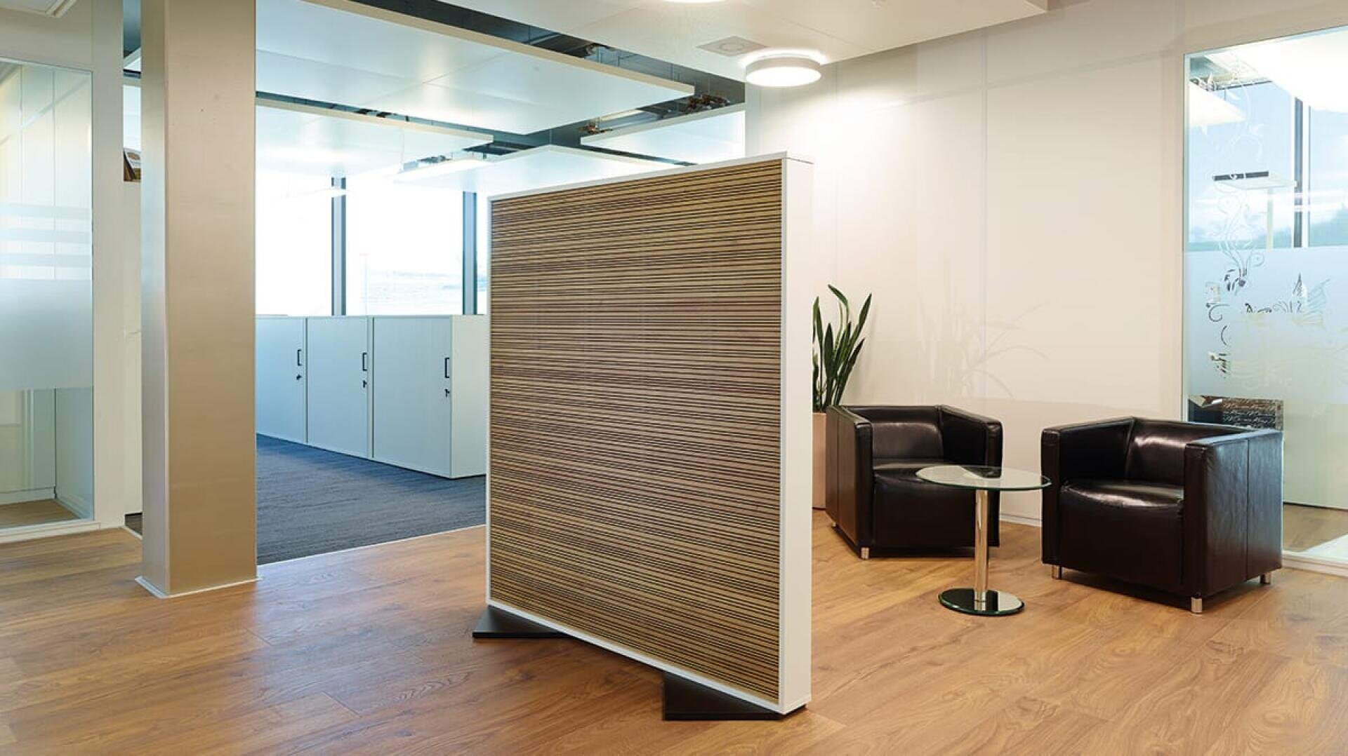 Ein moderner Büro-Wartebereich mit zwei schwarzen Sesseln, einem kleinen runden Glastisch, einer Pflanze und einer Holzwand auf einem hellen Holzboden. Im Hintergrund befinden sich Wände und Schränke aus Milchglas.