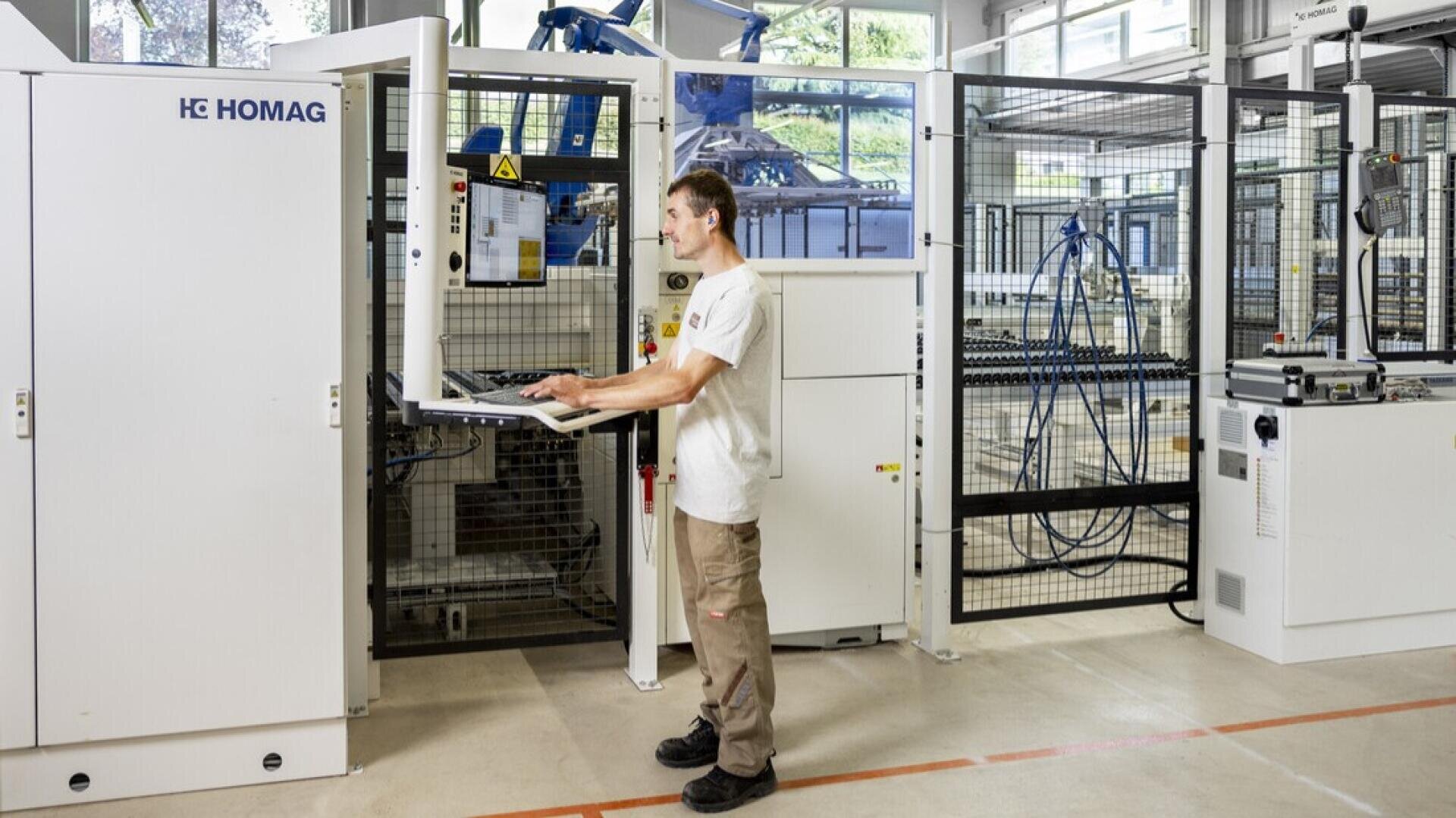 Un homme vêtu d'une chemise blanche et d'un pantalon kaki fait fonctionner une machine industrielle avec une interface informatique dans une usine moderne et lumineuse. Divers équipements et barrières de sécurité sont visibles à l'arrière-plan.