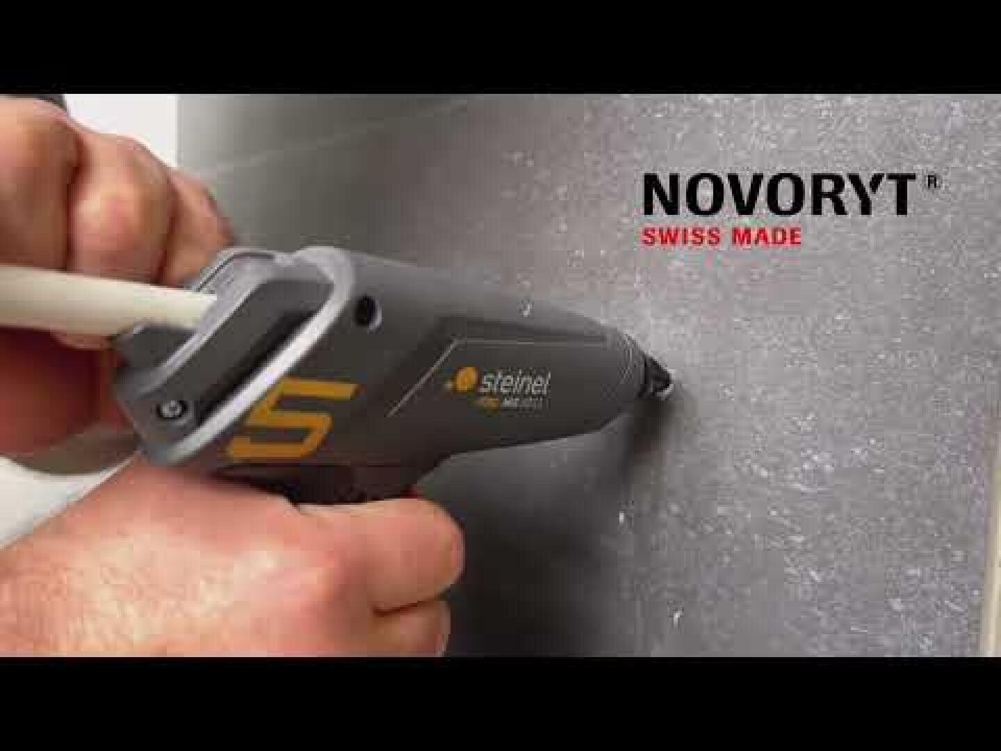 Une personne utilise un pistolet à colle Steinel gris pour remplir un trou dans un mur gris. Le texte NOVORYT SWISS MADE apparaît dans le coin supérieur droit.