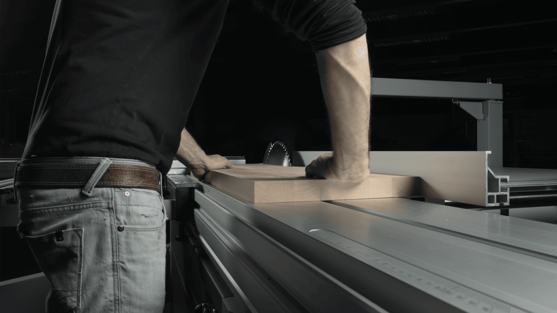 Eine Person, die ein schwarzes Hemd und eine graue Jeans trägt, schiebt in einer Werkstatt ein Holzstück durch eine Tischsäge, wobei die Hände das Holz zum Kreissägeblatt führen.