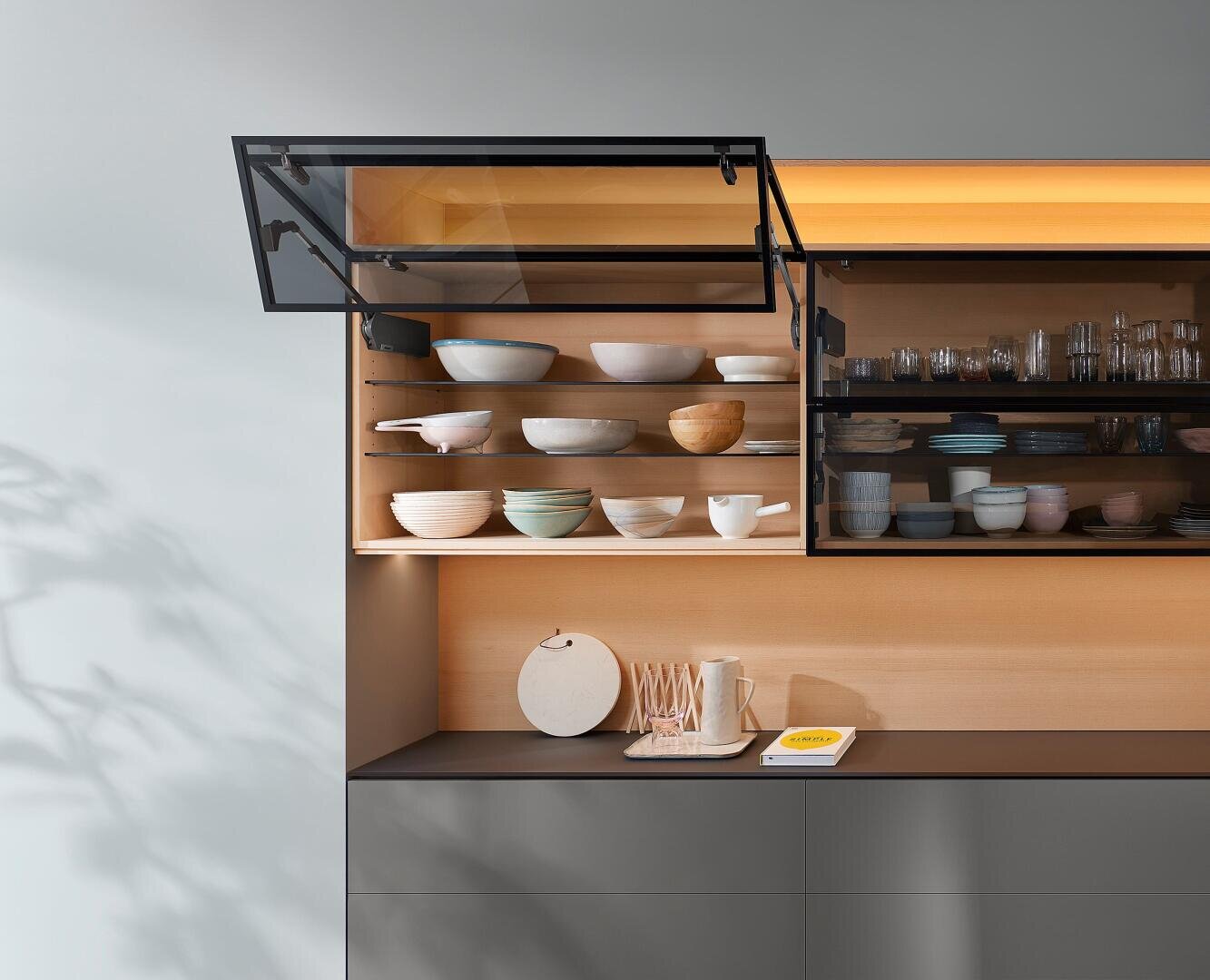 Ein moderner Küchenschrank mit einer nach oben geöffneten Glastür gibt den Blick auf ordentlich angeordnete Schüsseln und Schalen in Holzregalen frei. Darunter befinden sich auf einer Theke Teller, Tassen und ein gelbes Buch. Warmes Licht beleuchtet die Regale.