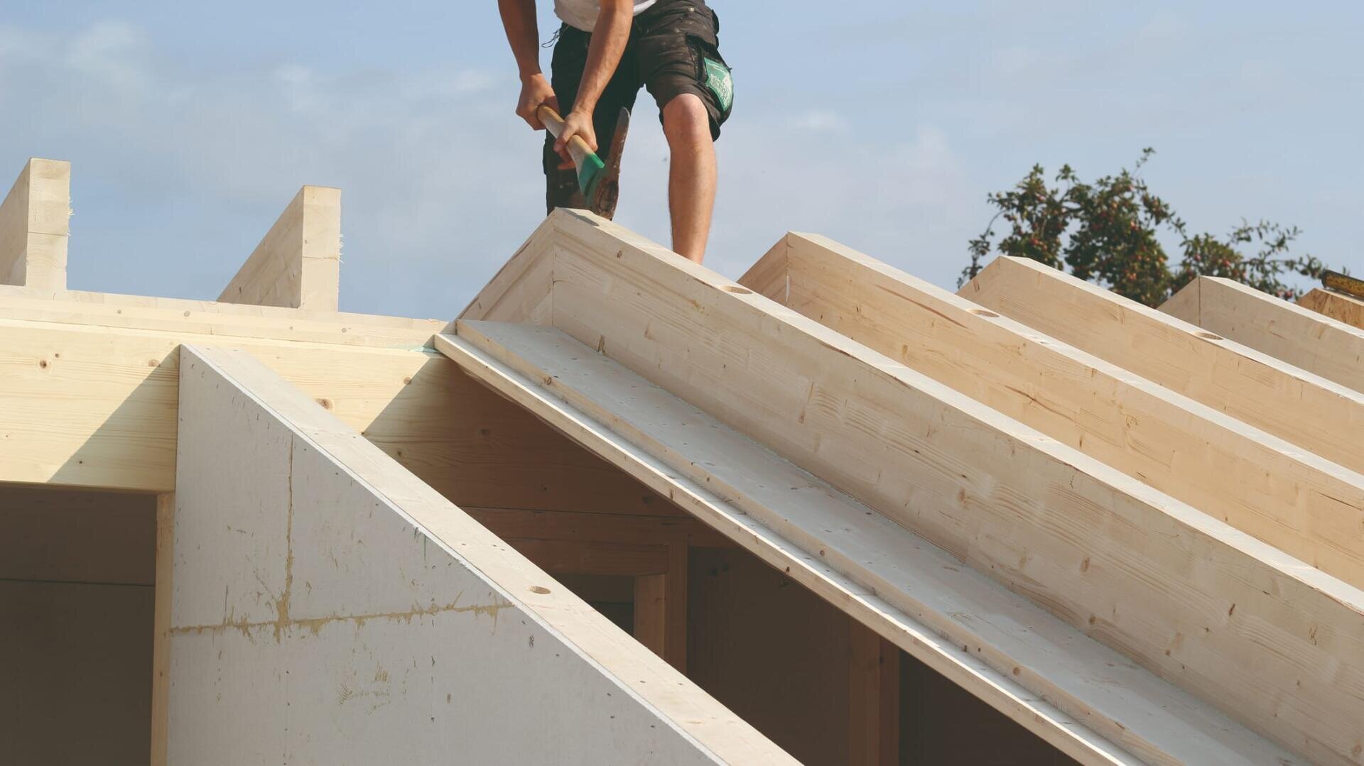 Eine Person steht auf dem Holzgerüst eines im Bau befindlichen Dachs und benutzt einen Hammer, um die Balken zu befestigen. Im Hintergrund sind der blaue Himmel und einige Bäume zu sehen.
