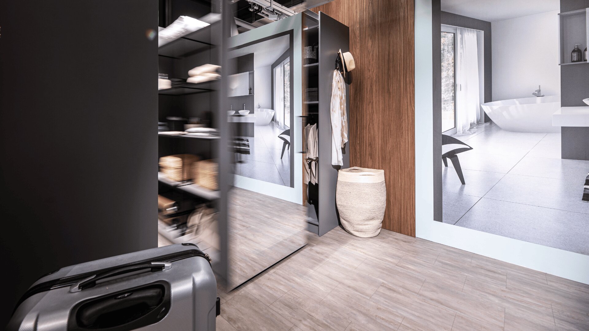 Un dressing moderne avec un grand miroir, des étagères ouvertes, des vêtements suspendus, une valise posée sur le sol et un panier à côté d'un mur lambrissé. Salle de bains bien éclairée avec une baignoire visible à travers une porte.