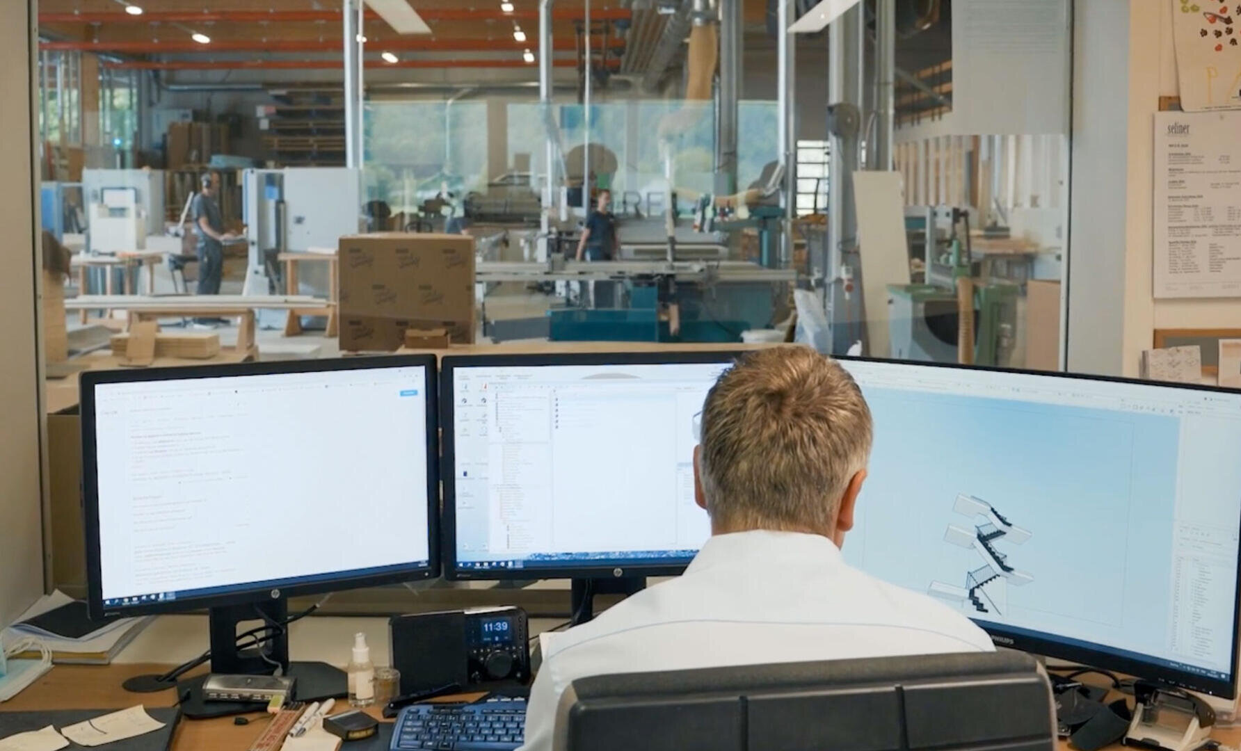 Eine Person sitzt an einem Schreibtisch mit drei großen Monitoren, auf denen technische Diagramme und Dokumente angezeigt werden, und blickt auf eine geräumige, moderne Industriewerkstatt mit verschiedenen Geräten und Arbeitern im Hintergrund.