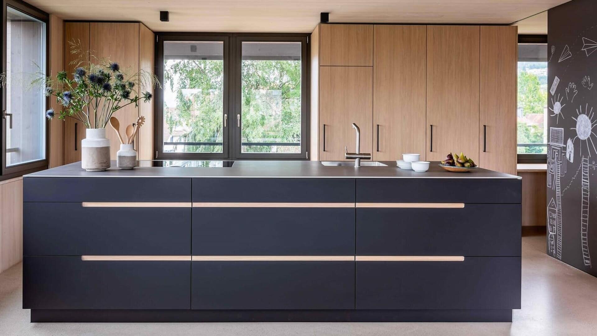 Cuisine moderne avec un grand îlot noir, des armoires en bois minimalistes, des vases décoratifs avec des fleurs, un bol de fruits et de grandes fenêtres laissant entrer la lumière naturelle. Un tableau noir avec des dessins est visible sur la droite.