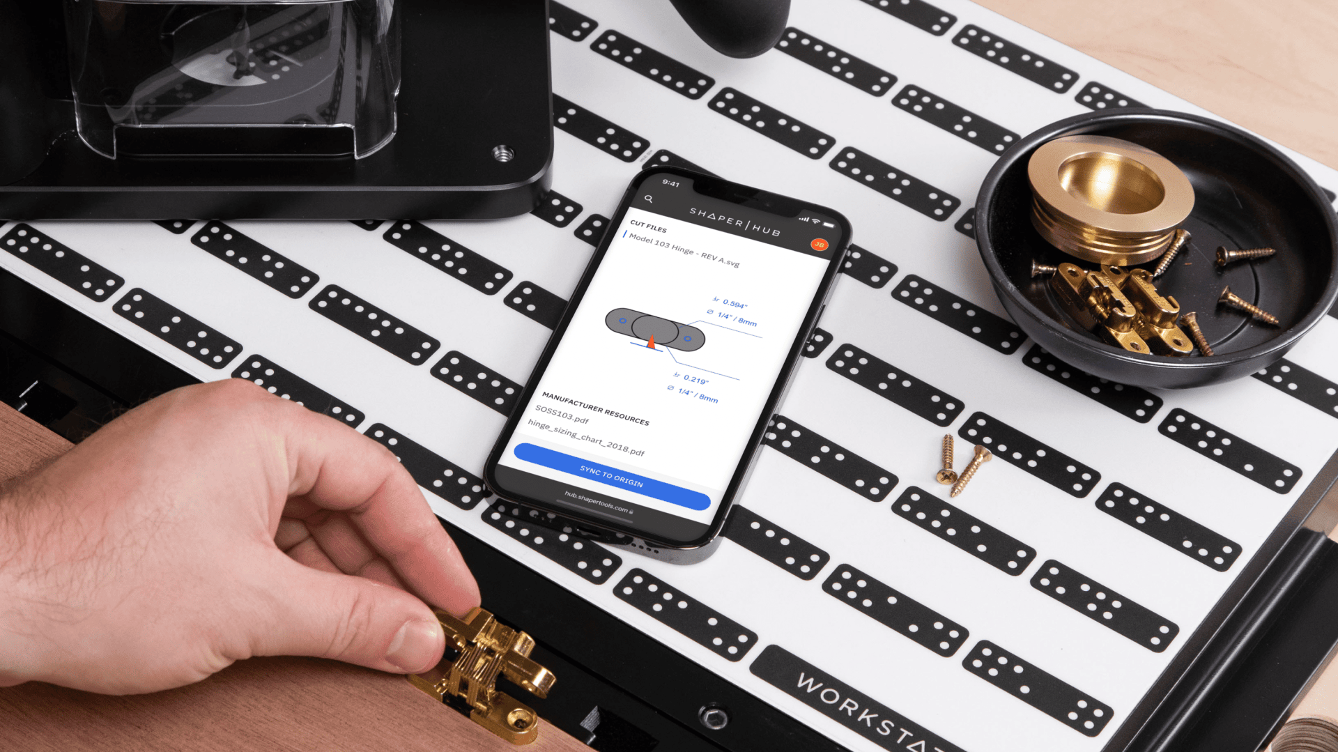Eine Person schraubt ein Scharnier an einer Werkbank fest, während sie ein Smartphone benutzt, das ein Diagramm und Maße für die genaue Platzierung anzeigt. Messingbeschläge und Schrauben liegen in einer kleinen schwarzen Schale daneben.