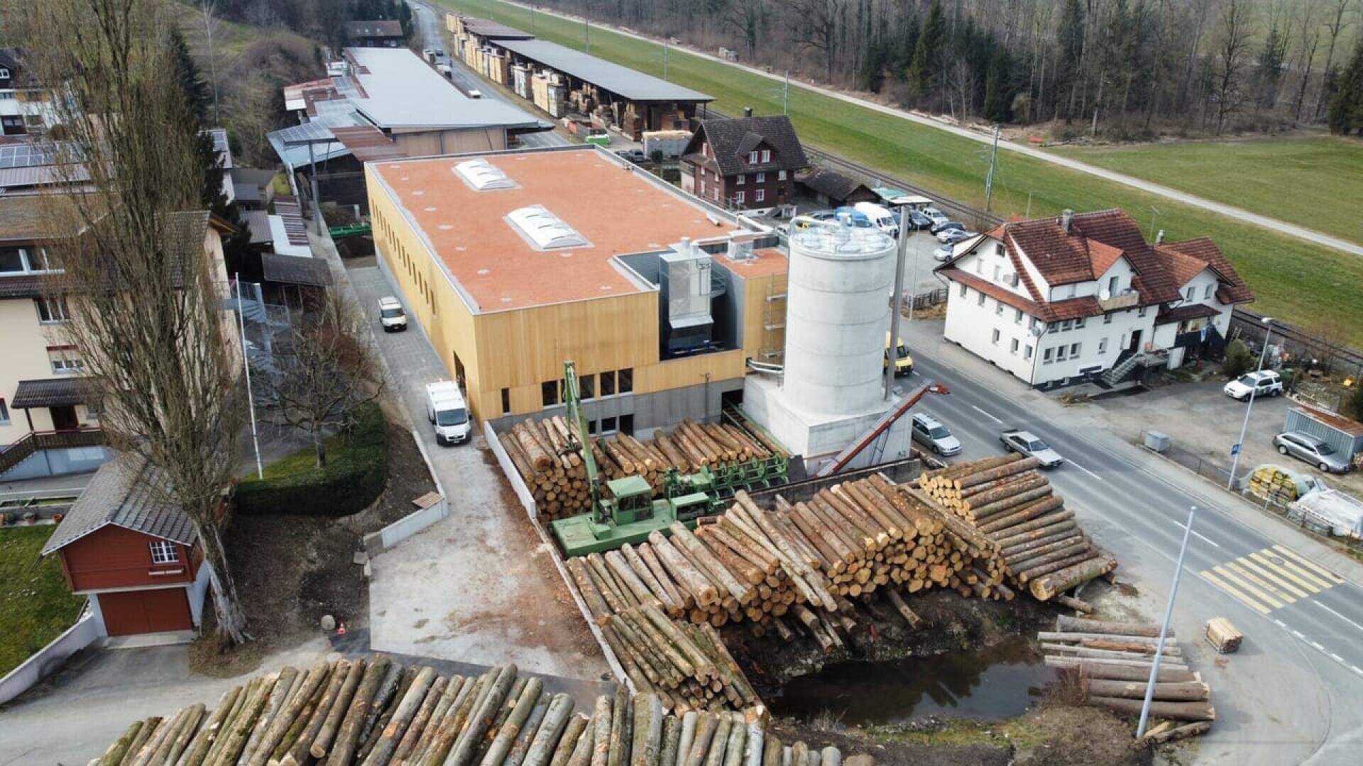 Vue aérienne d'un parc à bois avec de grandes piles de grumes, un bâtiment moderne avec un toit rouge, un grand silo blanc, et les maisons et routes environnantes dans une zone rurale.