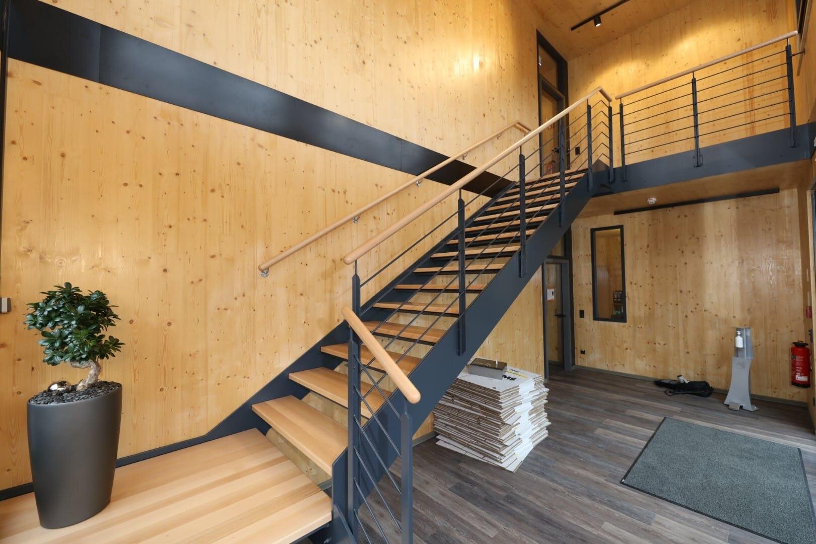 Modernes Interieur mit Holzwänden, einer schwarzen Metalltreppe mit Holzstufen und Geländer, einer Topfpflanze, gestapelten Holzpaneelen und grauem Bodenbelag. An der rechten Wand sind eine Tür und ein Fenster zu sehen.
