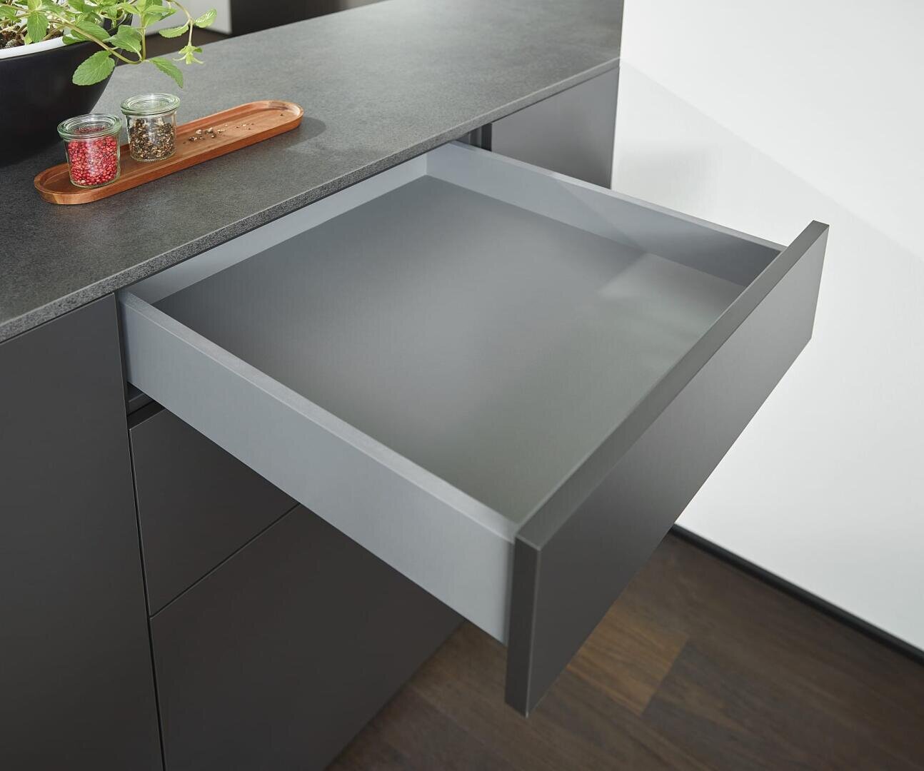 Eine moderne Küche mit einer schlanken, grauen Schublade, die aufgezogen ist, neben einer Arbeitsplatte mit einem Holztablett mit Gewürzdosen und einer kleinen Topfpflanze. Der Boden ist aus dunklem Holz.