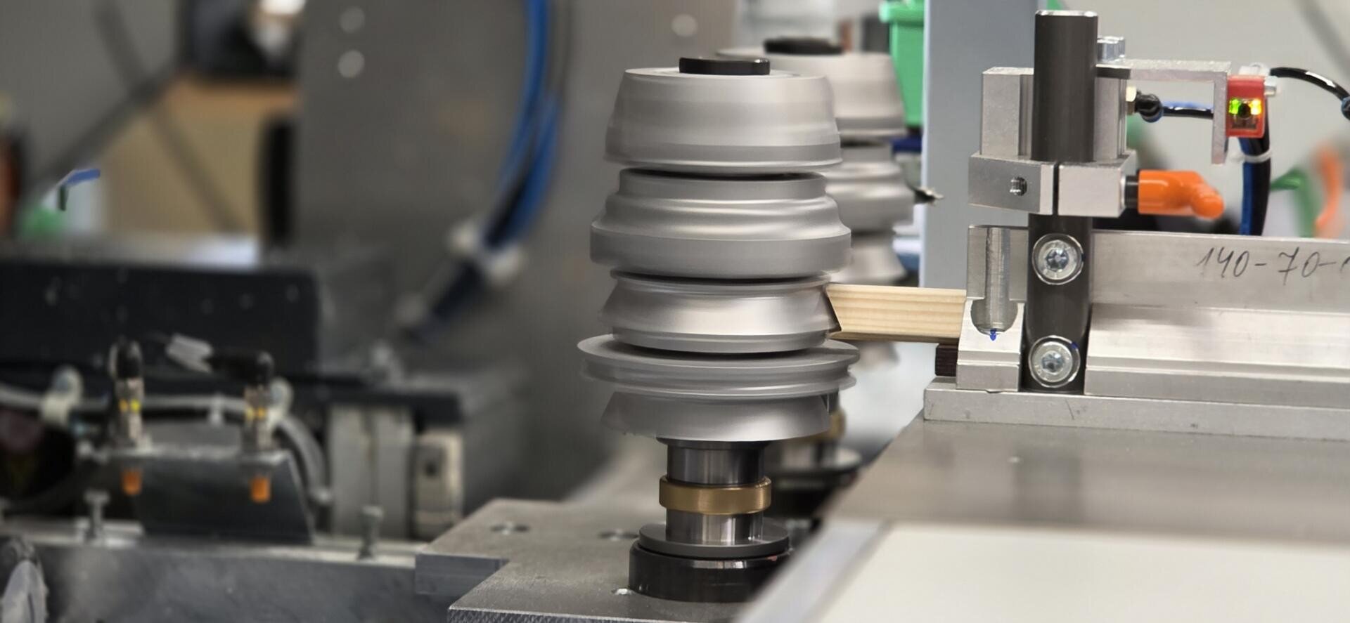 Nahaufnahme einer Industriemaschine, die einen Holzstab schleift oder poliert, mit rotierenden Metallrädern und verschiedenen mechanischen Komponenten in einer Fabrikumgebung.