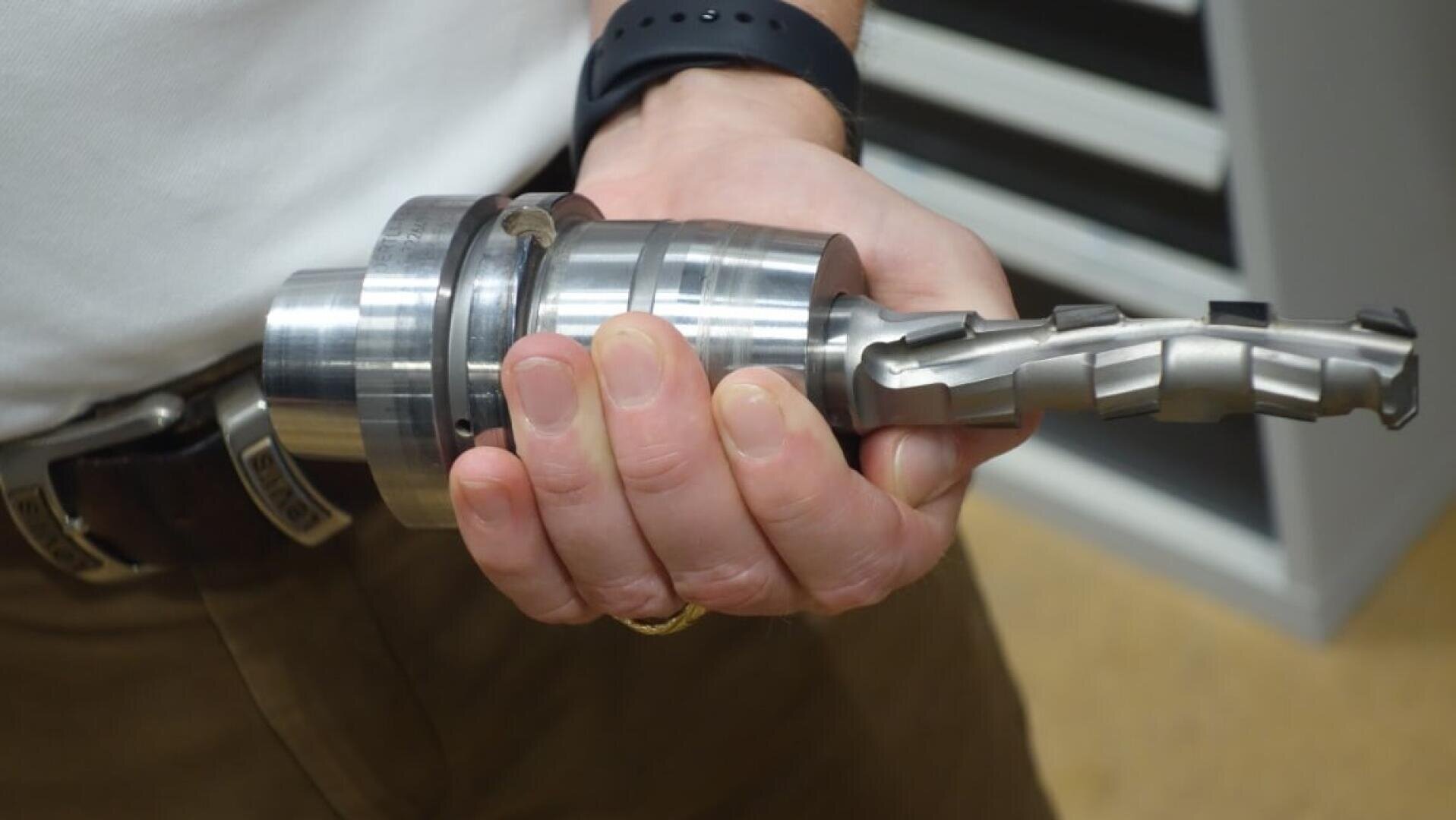 Eine Person hält ein großes industrielles Metallwerkzeug mit spiralförmiger Klinge und zylindrischer Basis, wahrscheinlich eine Bohrmaschine oder einen Fräser, in der rechten Hand. Die Person trägt ein weißes Hemd, eine braune Hose und eine schwarze Uhr.
