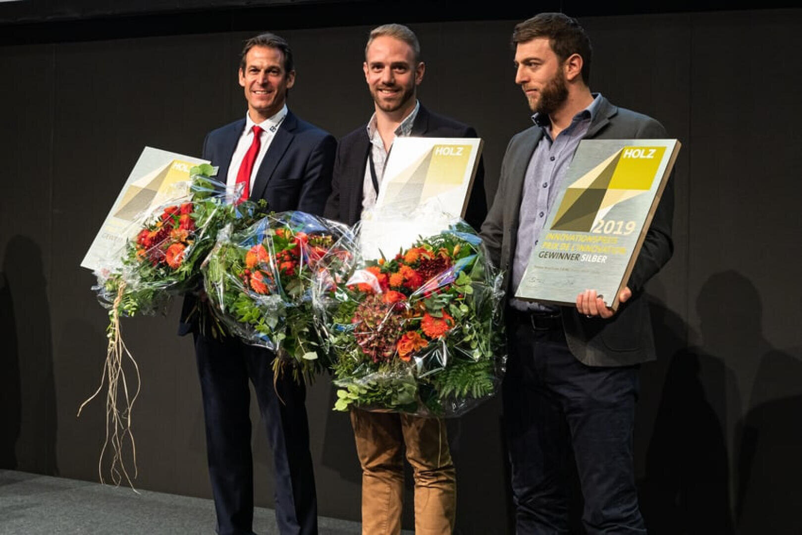 Drei Männer stehen nebeneinander, lächeln und halten große Blumensträuße und Urkunden in der Hand. Sie sind formell gekleidet, und der Hintergrund ist schlicht und dunkel.
