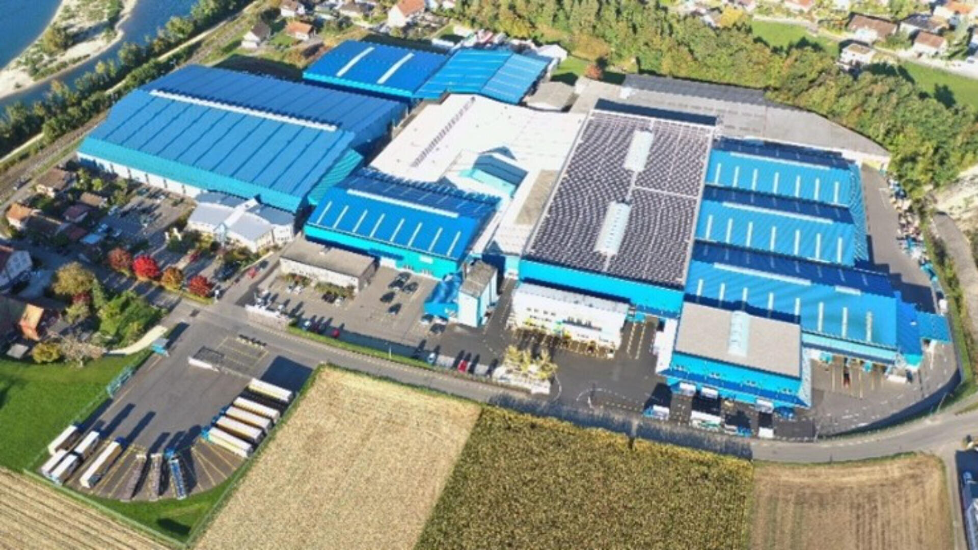 Luftaufnahme einer großen Industrieanlage mit mehreren Gebäuden mit blauen Dächern, umgeben von Feldern, Bäumen und Straßen, mit einem Wohngebiet in der Nähe. Auf einigen Dächern sind Sonnenkollektoren installiert.