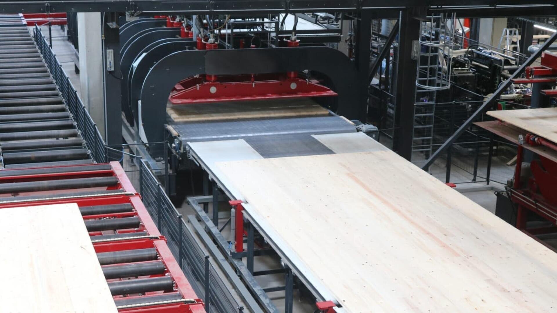 Dans une usine, une grande machine industrielle presse ou déplace un large panneau de bois le long d'un système de convoyage, entouré de cadres métalliques et de rouleaux.