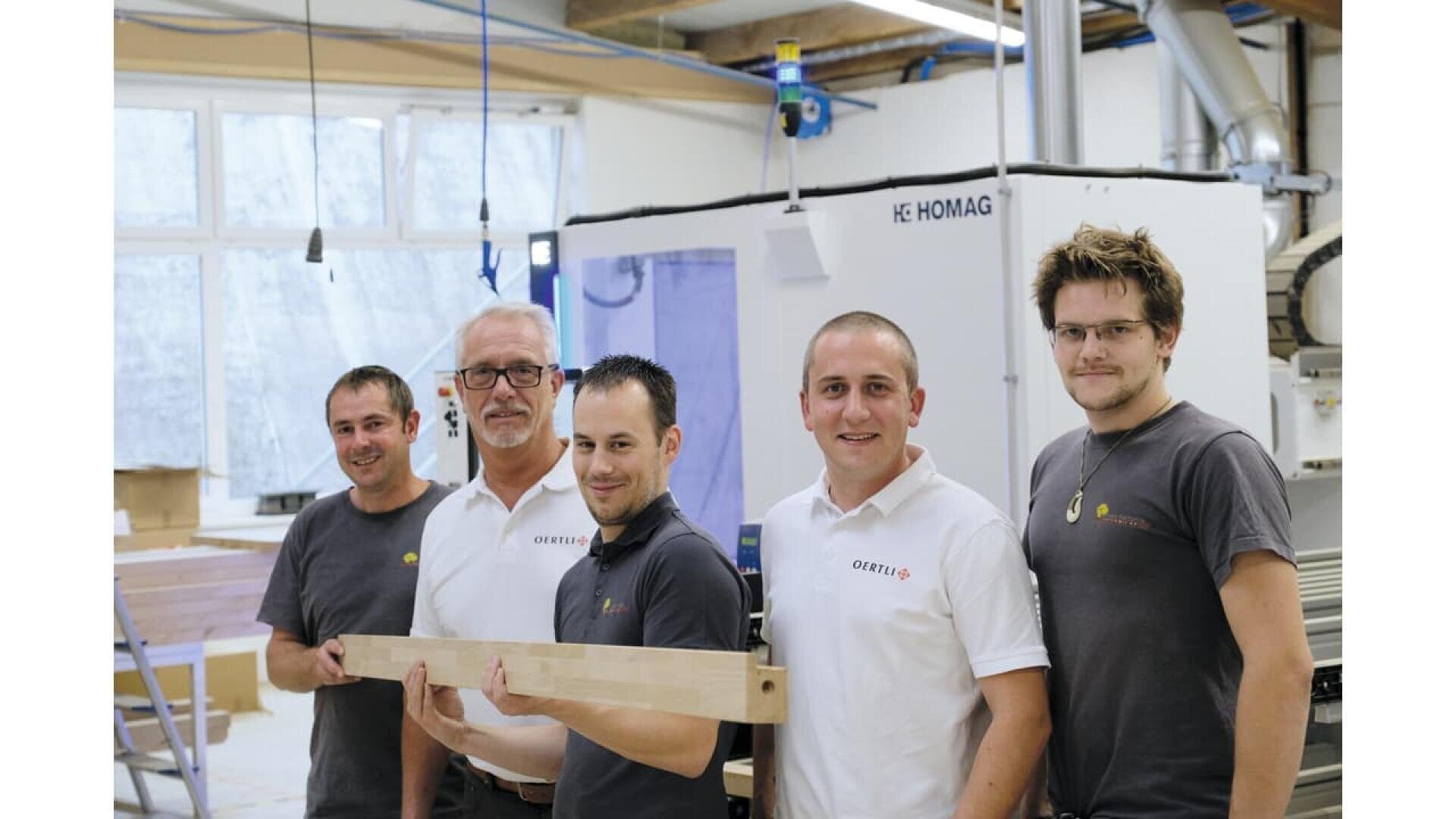 Cinq hommes portant des chemises de marque se tiennent dans un atelier et sourient à la caméra. Trois portent des chemises blanches, deux des chemises grises, et ils tiennent ensemble une planche de bois devant des machines à travailler le bois.