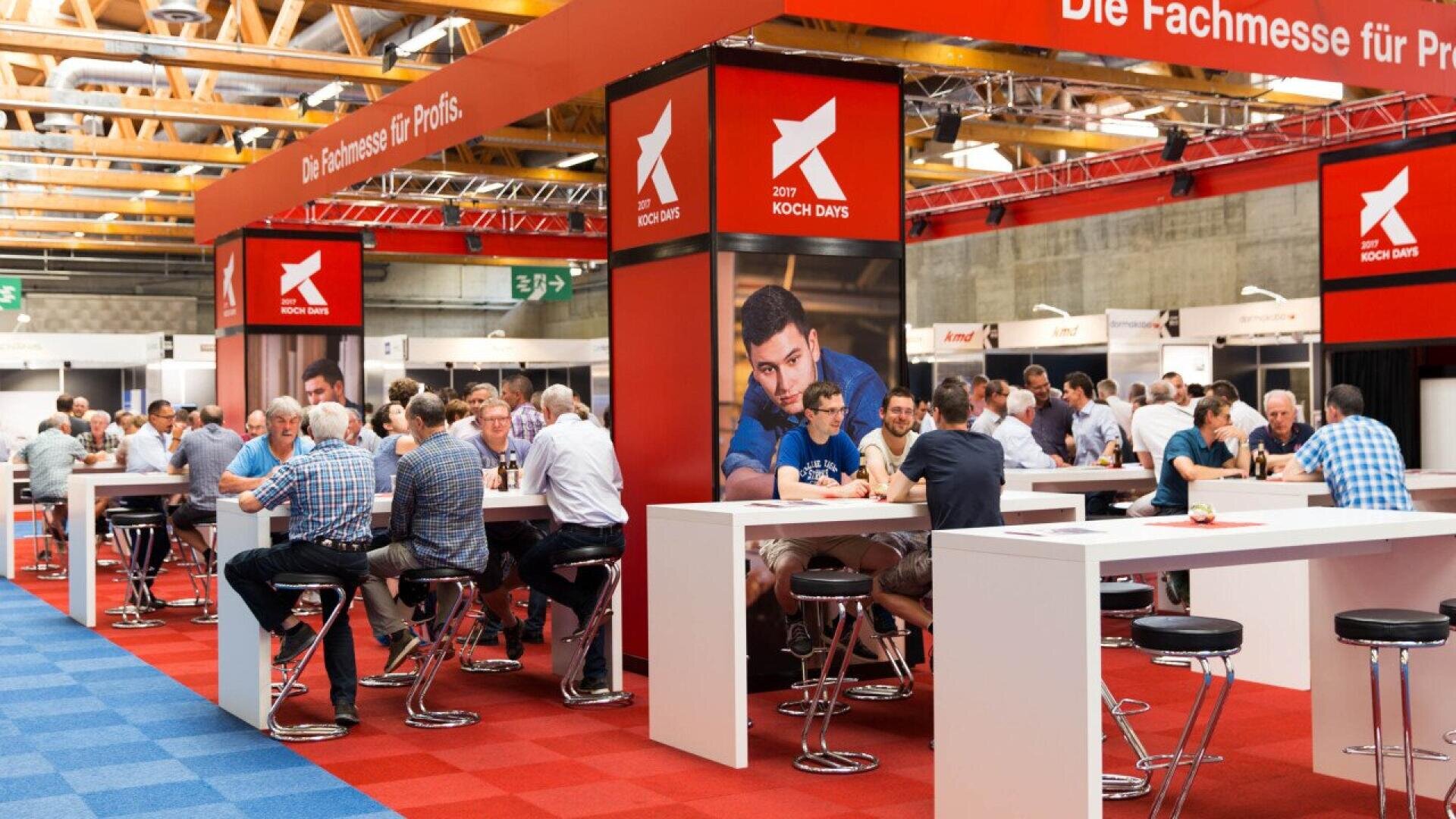 Des personnes sont assises à des tables hautes et à des tabourets dans un grand hall d'exposition lumineux recouvert d'une moquette rouge et bleue. Des banderoles indiquant "KOCH DAYS" sont suspendues au-dessus de la salle, et l'atmosphère est animée et sociale.