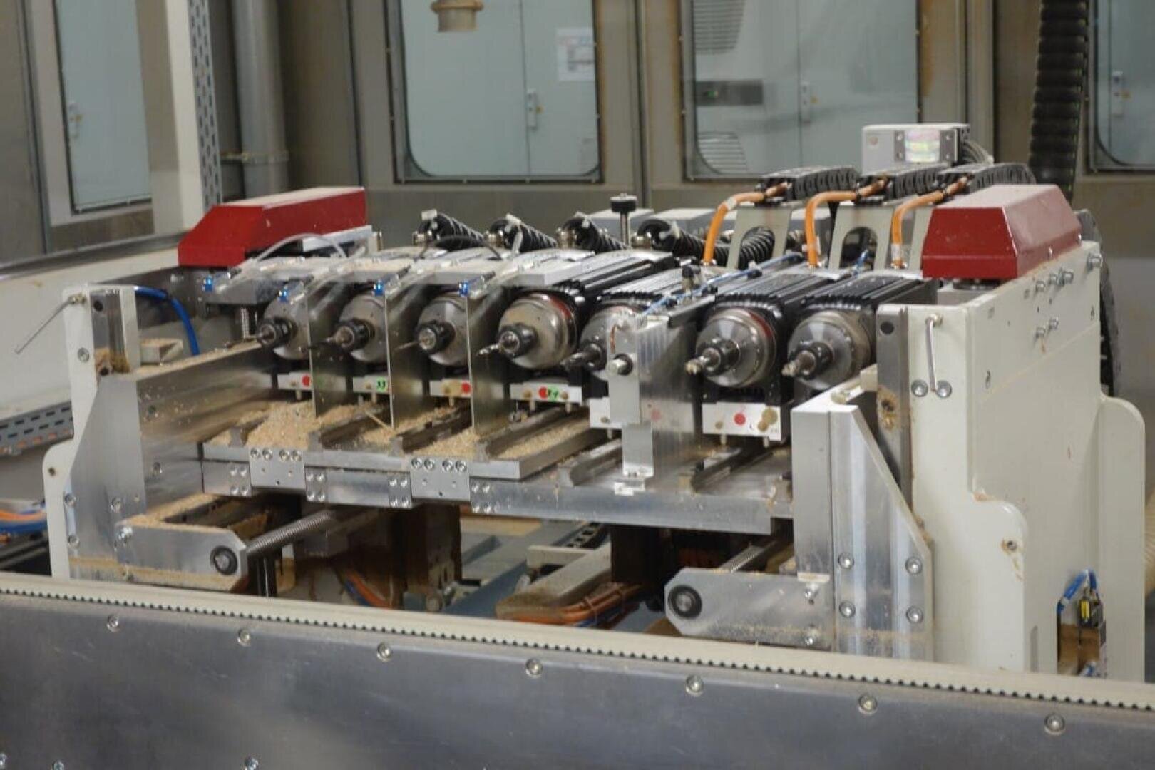 Eine Nahaufnahme einer industriellen Holzbearbeitungsmaschine mit mehreren Bohrköpfen und Motoren, umgeben von Holzspänen, in einer Produktionsstätte.