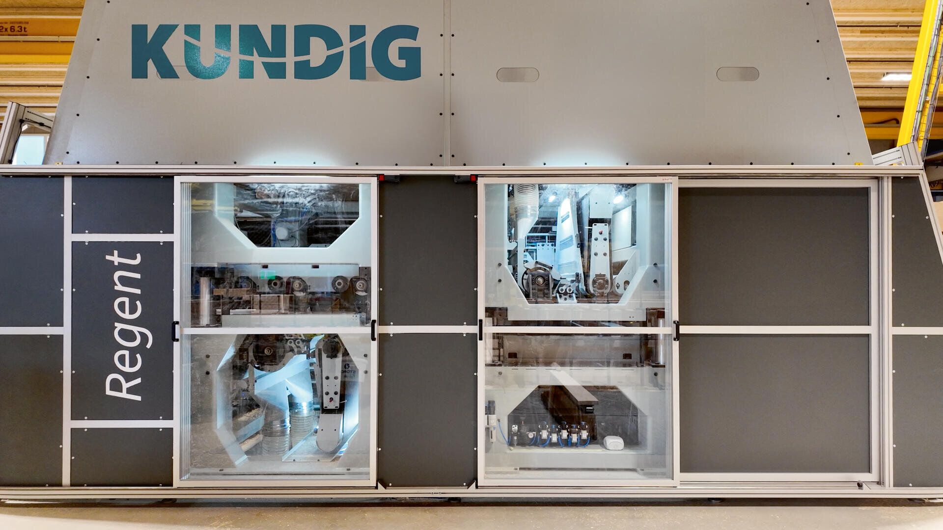 Eine große Industriemaschine mit der Bezeichnung Kundig Regent verfügt über drei transparente Paneele, die komplexe interne Mechanismen und Komponenten offenbaren. Das Äußere ist grau, und der Markenname ist oben gut sichtbar angebracht.