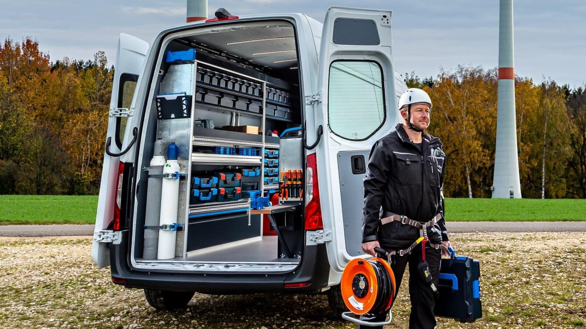 Un homme en tenue de travail et portant un casque se tient à côté d'une camionnette ouverte remplie d'outils et d'équipements organisés, tenant un enrouleur de câble et une boîte à outils. La scène se déroule en extérieur, près d'éoliennes et d'un champ herbeux.