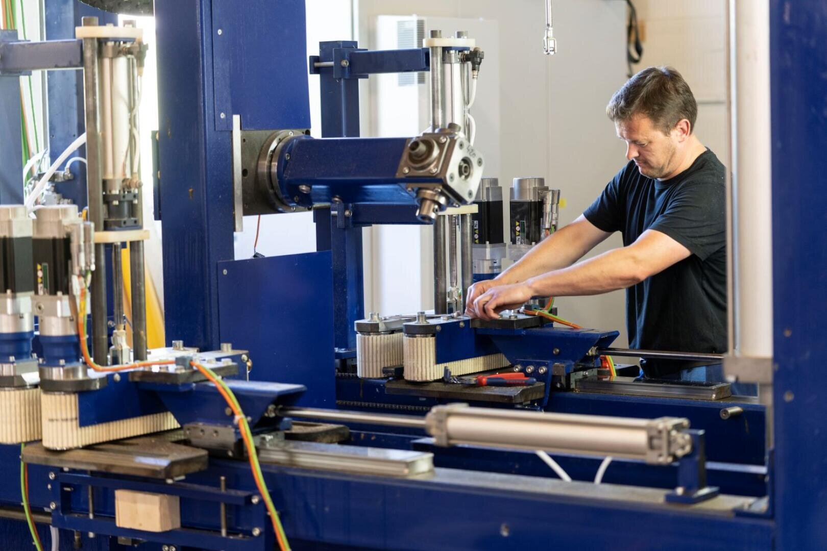 Ein Mann mit einem schwarzen Hemd bedient in einer Fabrik oder Werkstatt eine große blaue Industriemaschine mit vielen Metallteilen, Drähten und Kolben.