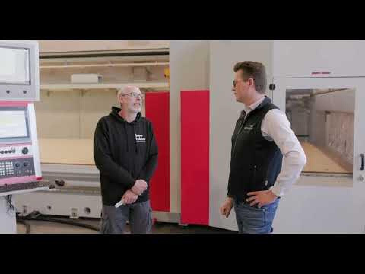 Zwei Männer stehen und unterhalten sich in einer Industrieumgebung in der Nähe großer Maschinen und Schalttafeln. Einer trägt einen schwarzen Kapuzenpulli und eine Brille, der andere ein weißes Hemd mit einer schwarzen Weste.