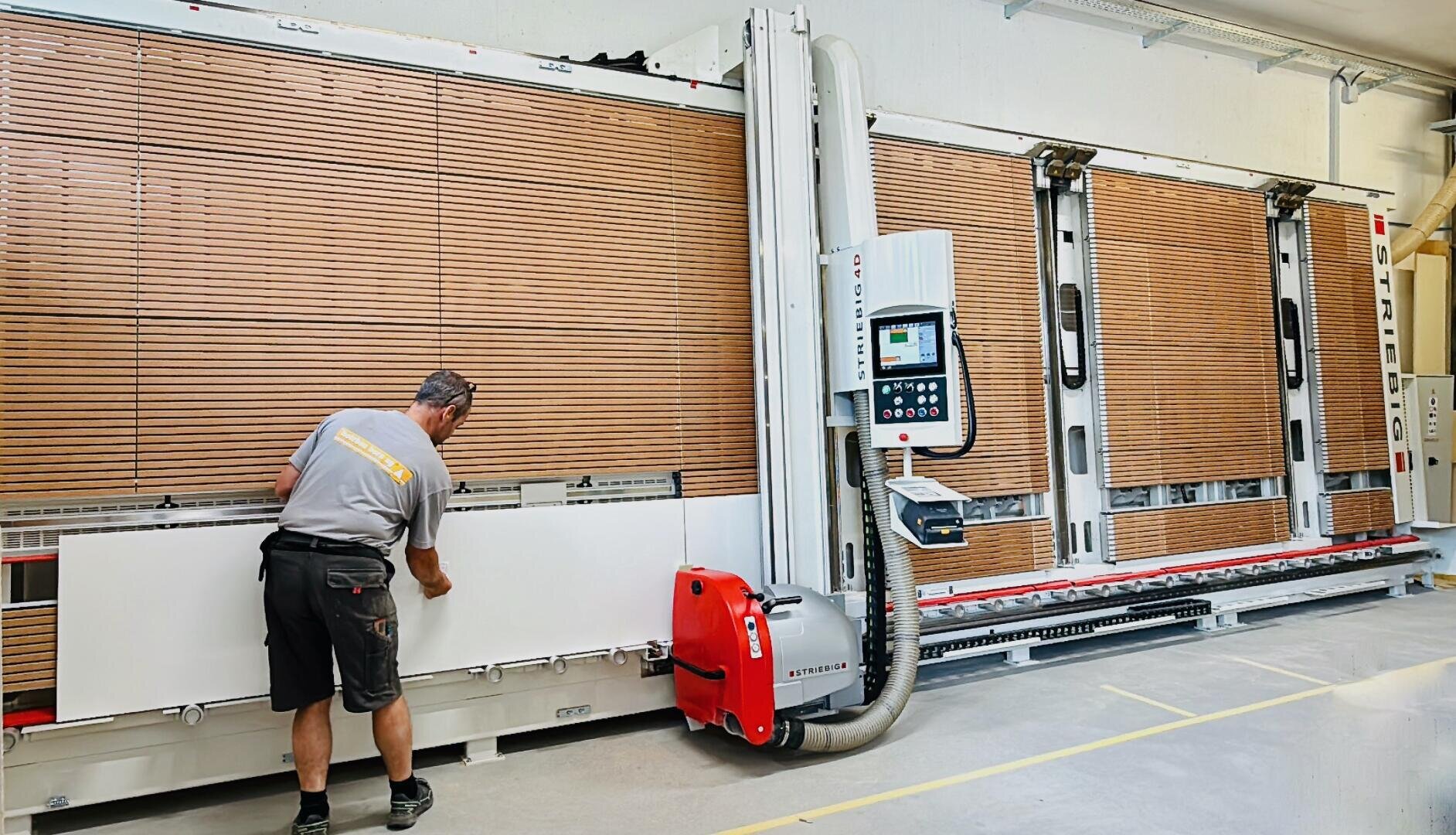 Un homme utilise une grande scie à panneaux verticale dans un atelier et découpe un tableau blanc. La machine a un panneau de commande, des dispositifs de sécurité et des lattes de bois en arrière-plan. Le sol est marqué par des lignes de sécurité jaunes.