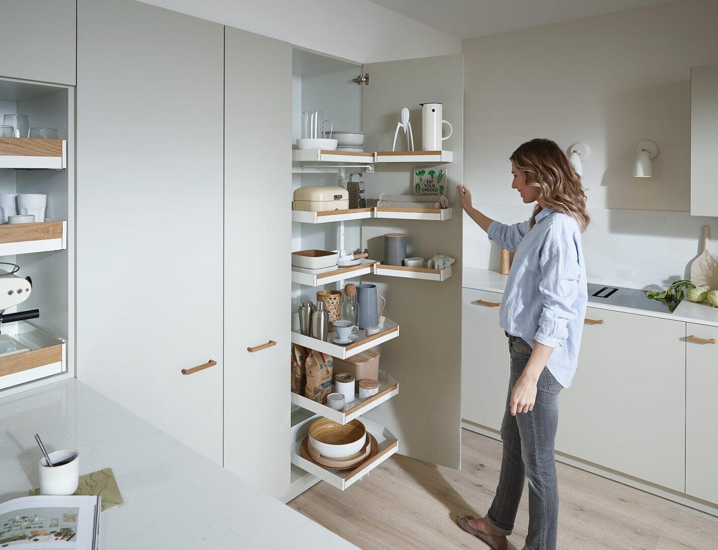 Eine Frau steht in einer modernen Küche und greift in einen Eckschrank mit drehbaren Regalen, die ordentlich mit Geschirr, Schüsseln, Gläsern und Küchenutensilien bestückt sind. Der Raum ist hell und minimalistisch mit hellen Schränken.