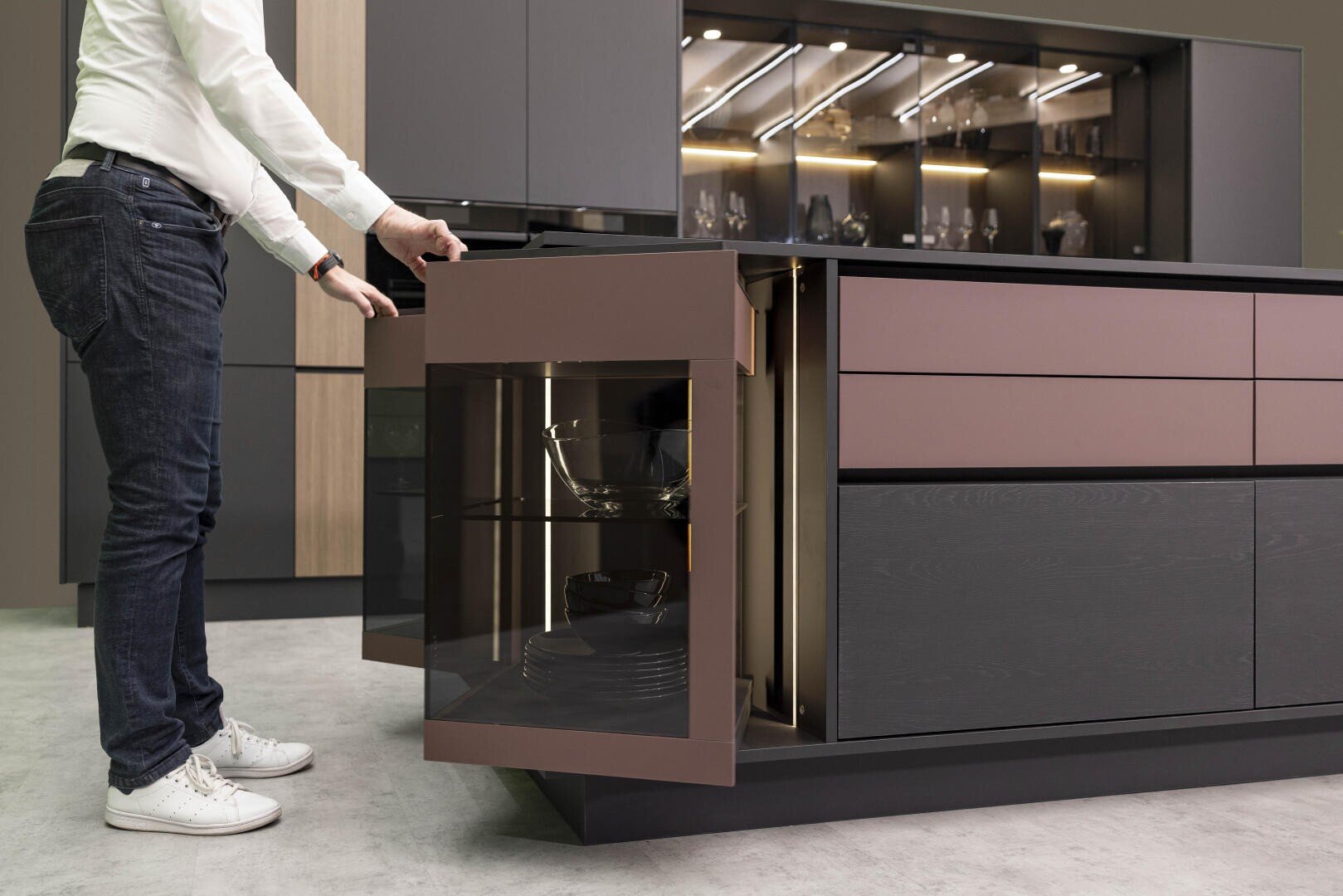 Eine Person öffnet eine Eckvitrine mit Glasfront in einer modernen Küche mit schlichten, dunkelbraunen und schwarzen Schränken und gibt den Blick frei auf Regale mit Schüsseln und Geschirr darin.
