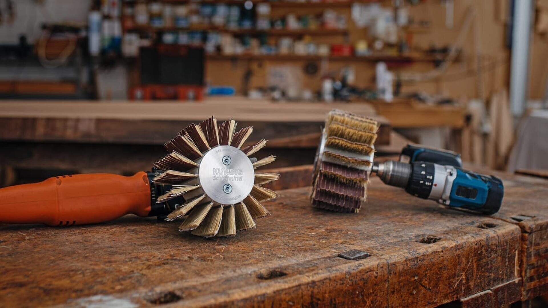 Gros plan sur un établi avec deux outils à brosse rotative, l'un avec un manche orange et l'autre fixé à une perceuse sans fil. Les étagères contenant des outils et des matériaux sont floues à l'arrière-plan.