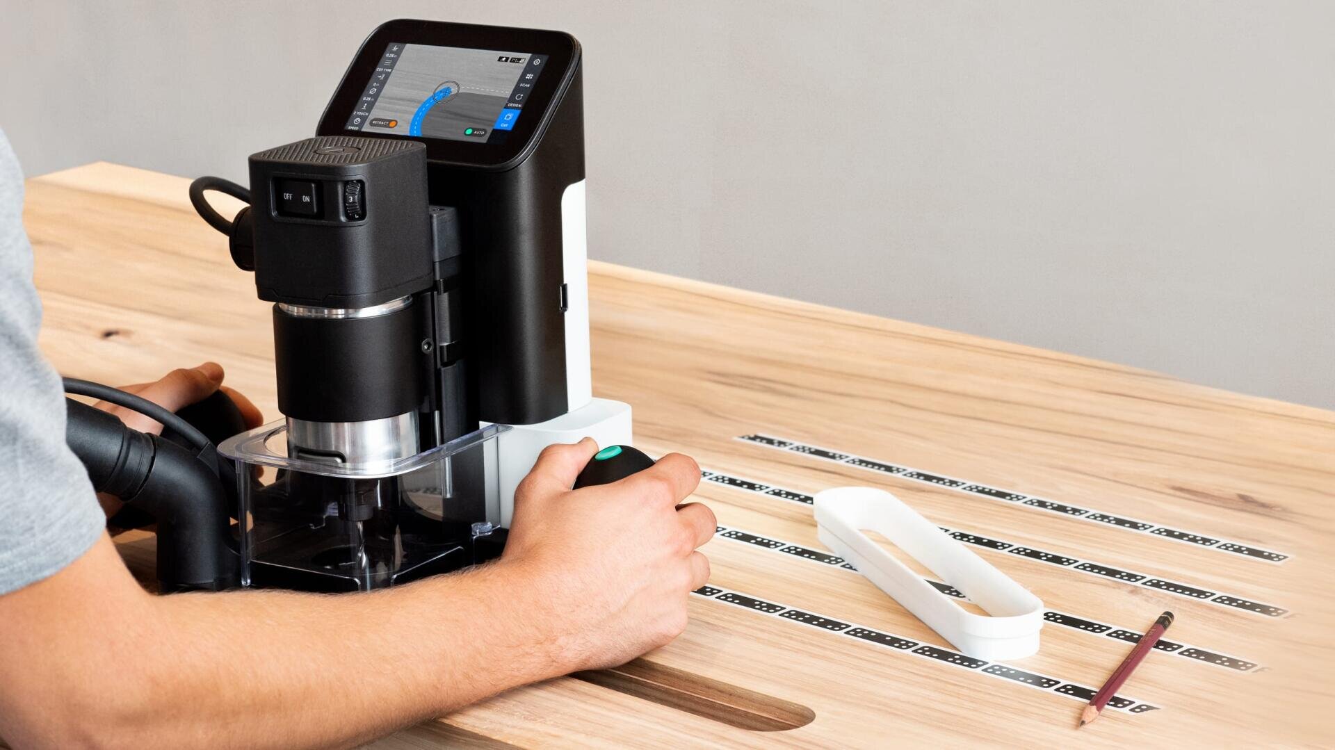 Eine Person bedient eine handgeführte CNC-Fräse mit digitalem Bildschirm und führt sie entlang von Strichcode-Streifen auf einer Holzoberfläche. Ein Bleistift und eine weiße Schablone liegen in der Nähe auf dem Tisch.