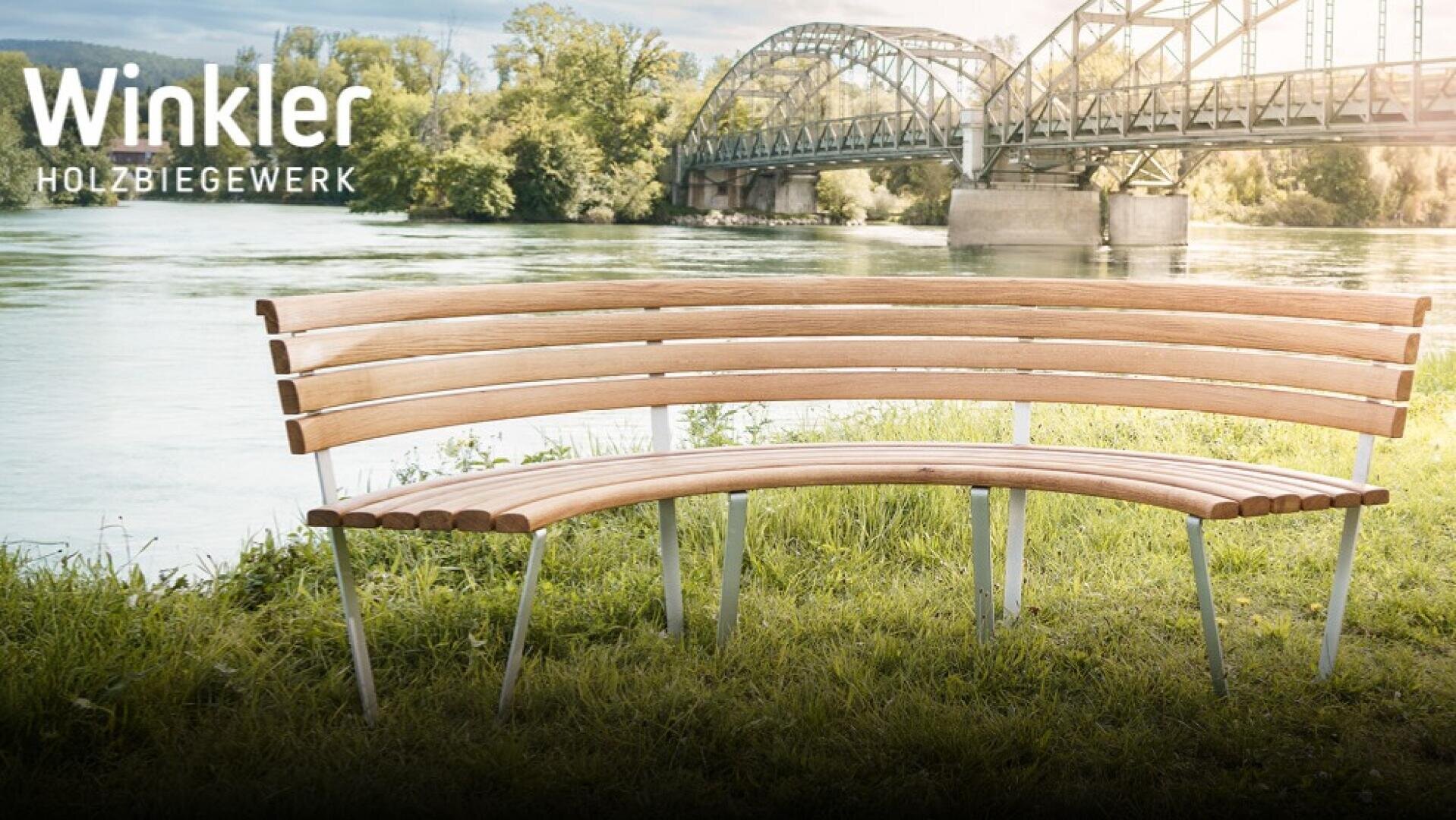 Banc en bois incurvé avec des pieds en métal sur une rive herbeuse, avec un pont métallique et des arbres à l'arrière-plan. Le texte indique "Winkler HOLZBIEGEWERK" dans le coin supérieur gauche.