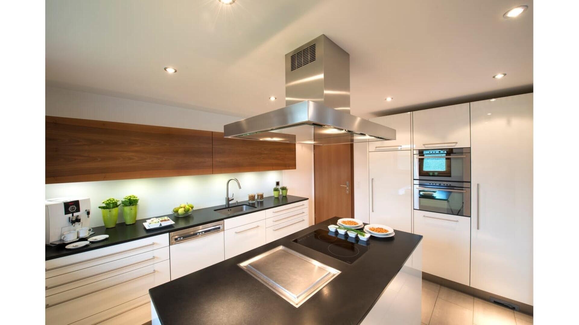 Cuisine moderne avec des armoires blanches, des comptoirs noirs, des appareils encastrés, un îlot avec une table de cuisson et une hotte en acier inoxydable. Des plantes en pot et des assiettes de nourriture ajoutent des touches décoratives.