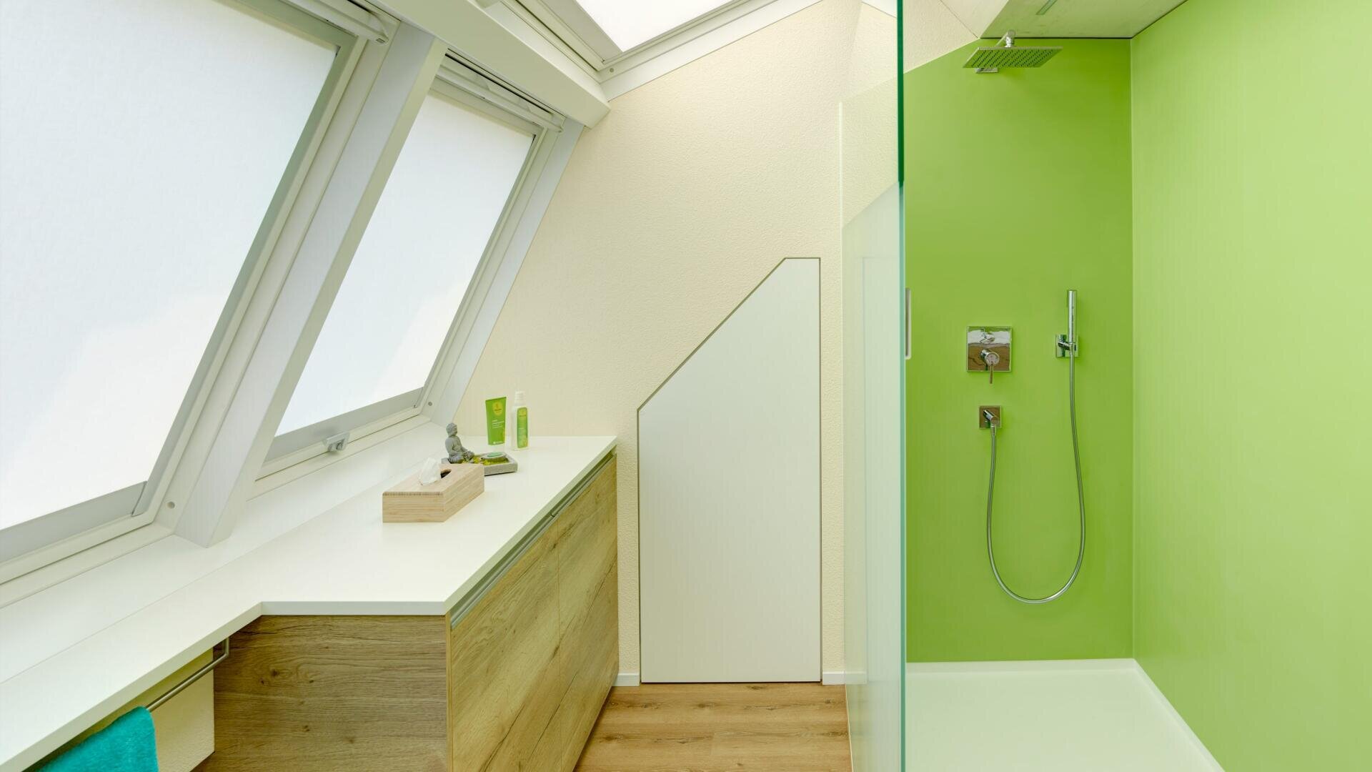 Ein modernes Badezimmer mit schräger Decke, großen Dachfenstern, Holzschränken, weißer Arbeitsplatte und einer gläsernen Dusche mit hellgrünen Wänden und einer Regendusche.