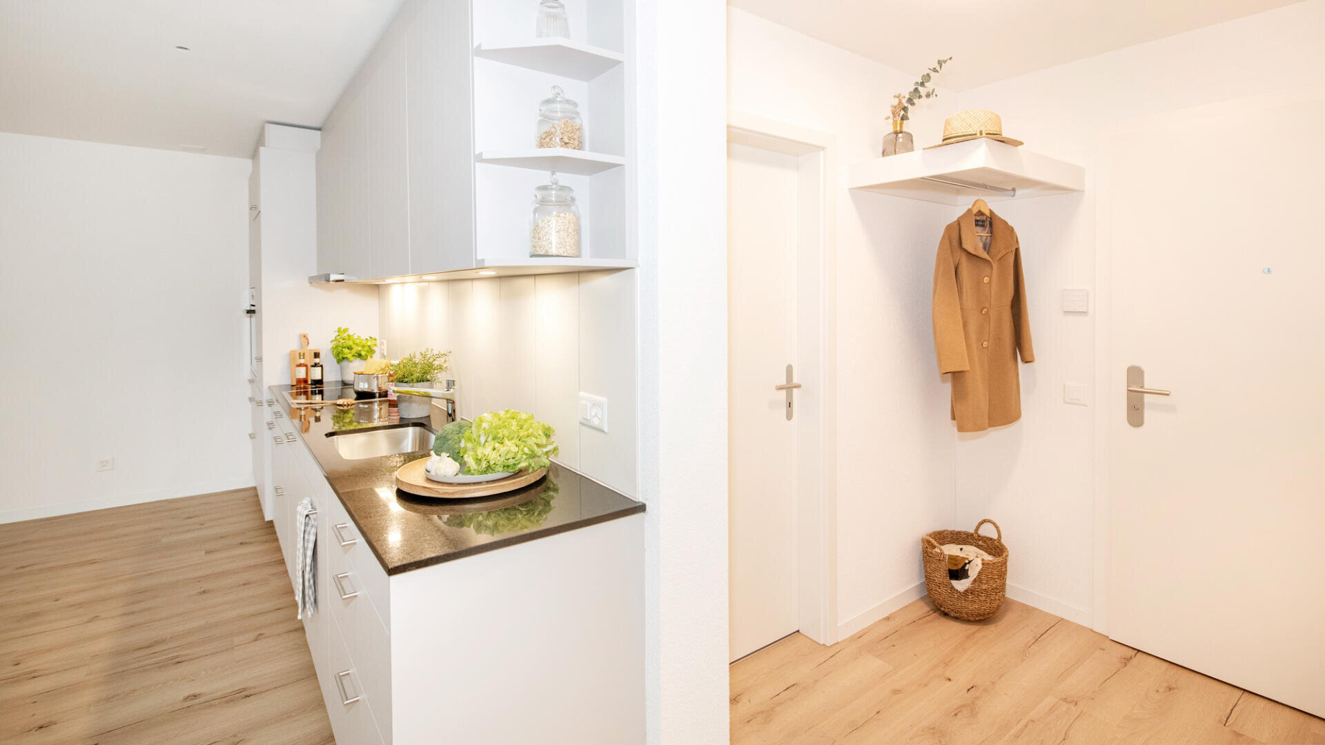 Eine moderne, minimalistische Küche mit weißen Schränken und Holzböden, angrenzend an einen hellen Eingangsbereich mit einem Garderobenständer mit einem hellbraunen Mantel, einem Strohhut und einem geflochtenen Korb auf dem Boden.