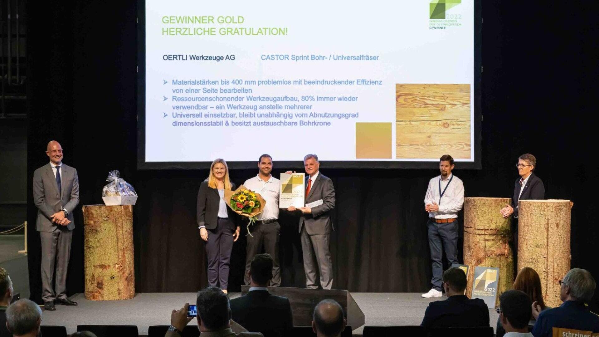 Un groupe de cinq personnes se tient sur une scène lors d'une cérémonie de remise de prix. Une personne tient un certificat et une autre un bouquet. Derrière eux, une diapositive de présentation félicite la société OERTLI Werkzeuge AG pour avoir remporté un prix d'or.