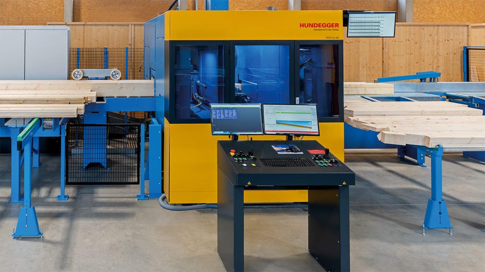 Eine gelbe und blaue Hundegger-Industrieholzbearbeitungsmaschine mit einem Computersteuerungsplatz davor, umgeben von Holzbrettern in einer Werkstattumgebung.