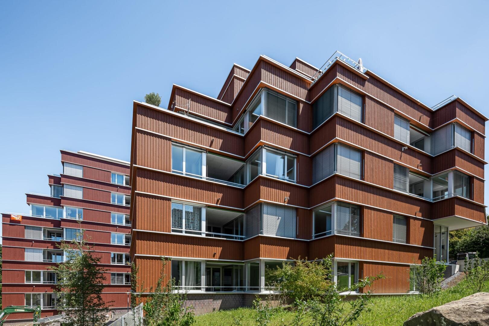 Moderne Mehrfamilienhäuser mit winkelförmigen, sich überlappenden Balkonen und großen Fenstern, mit rotbraunen Fassaden, unter einem klaren blauen Himmel, umgeben von grüner Vegetation.