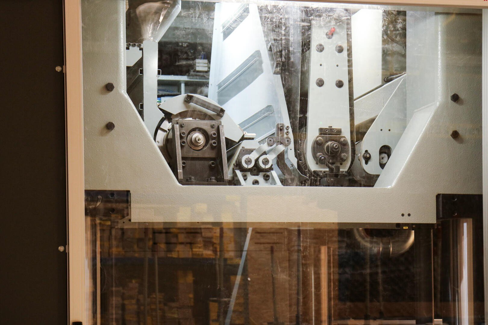 Nahaufnahme einer Industriemaschine mit Metallteilen und Zahnrädern hinter einer schützenden Glasscheibe in einer Fabrik oder einer Produktionsumgebung.