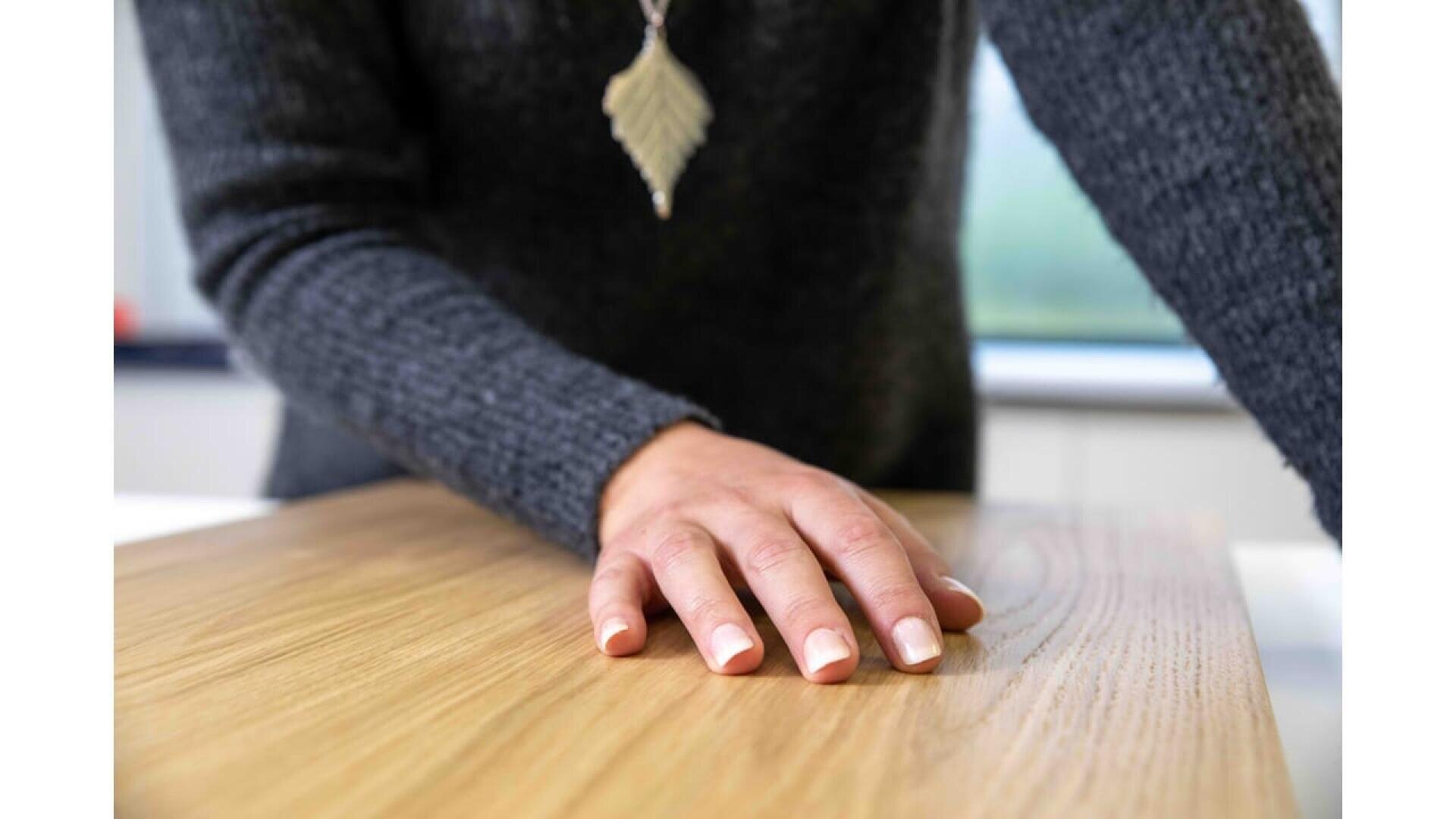 Une personne portant un pull gris et un collier en forme de feuille pose sa main sur une table en bois, avec en arrière-plan une lumière naturelle provenant d'une fenêtre.