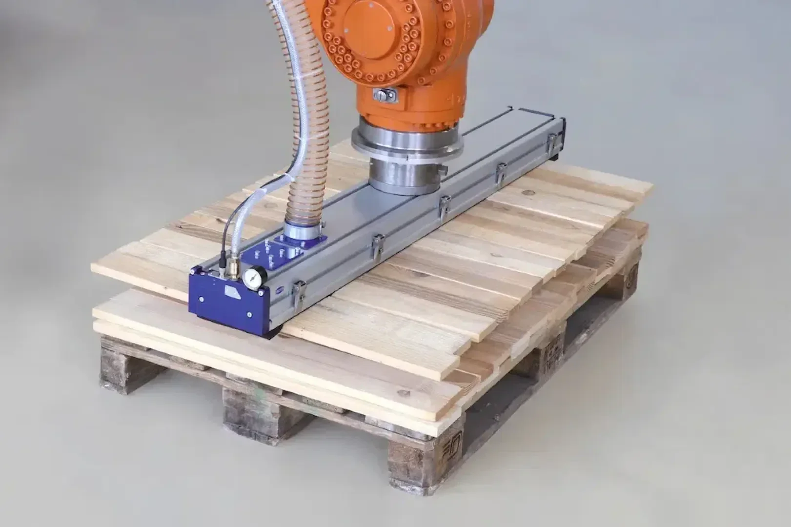 Ein Roboterarm mit einer Saugvorrichtung hebt oder legt Holzbretter auf eine Holzpalette auf einem glatten, hellen Boden.