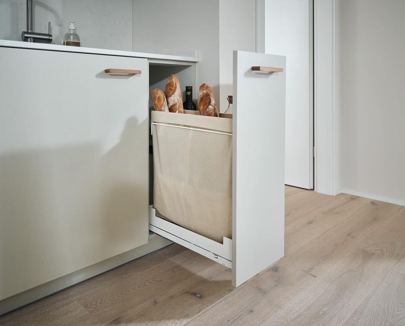 Eine moderne Küche mit einer ausziehbaren Vorratsschublade, die teilweise geöffnet ist und in der Brot und Flaschen aufbewahrt werden. Die Schublade ist in helle Schränke auf einem Holzboden integriert.