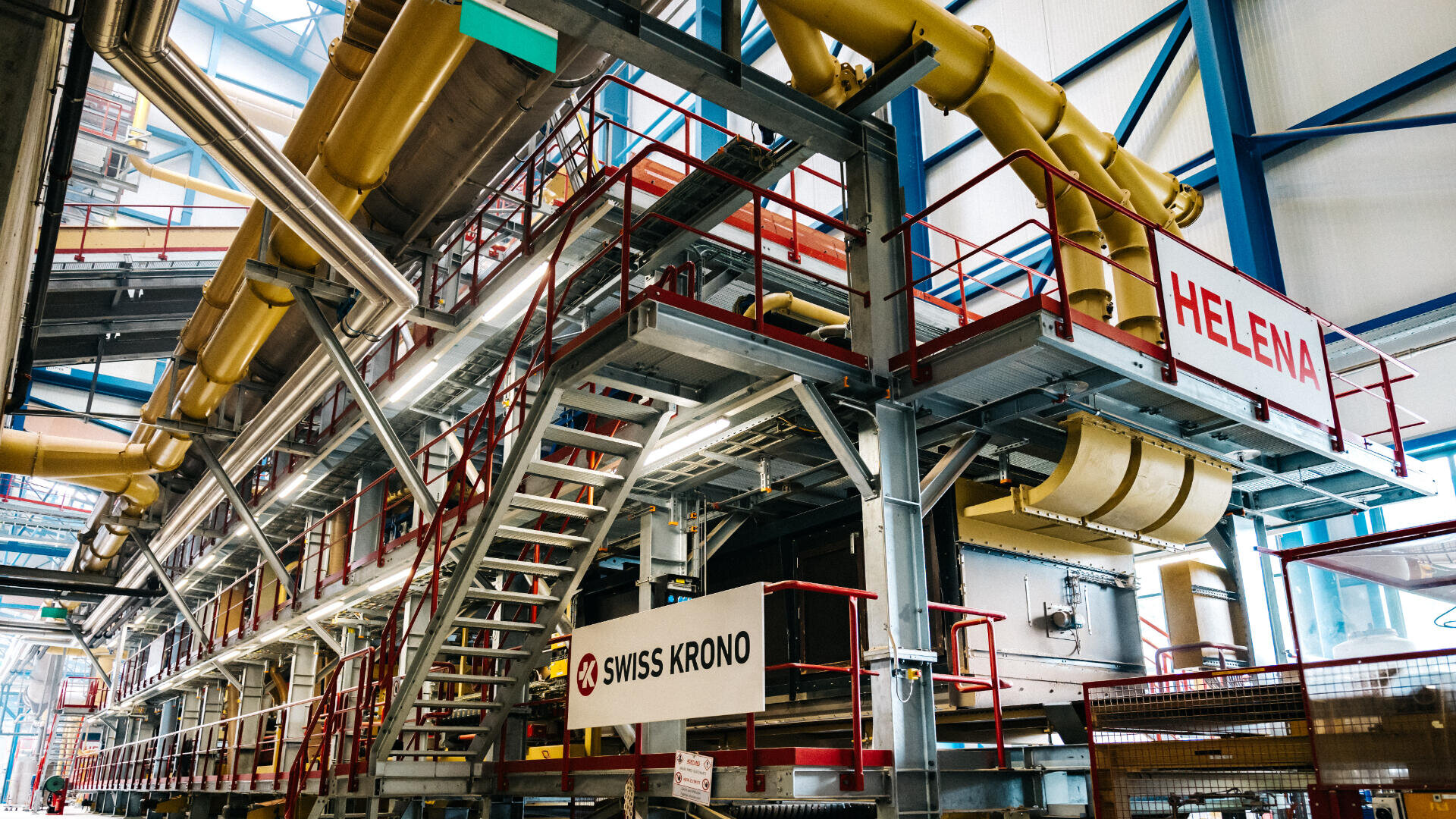 Industrielles Interieur mit großen Maschinen, Metalltreppen und Laufstegen. Auf den Schildern steht SWISS KRONO und HELENA. Gelbe Rohre und Balken verlaufen in einer geräumigen, hell erleuchteten Fabrikumgebung über Kopf.
