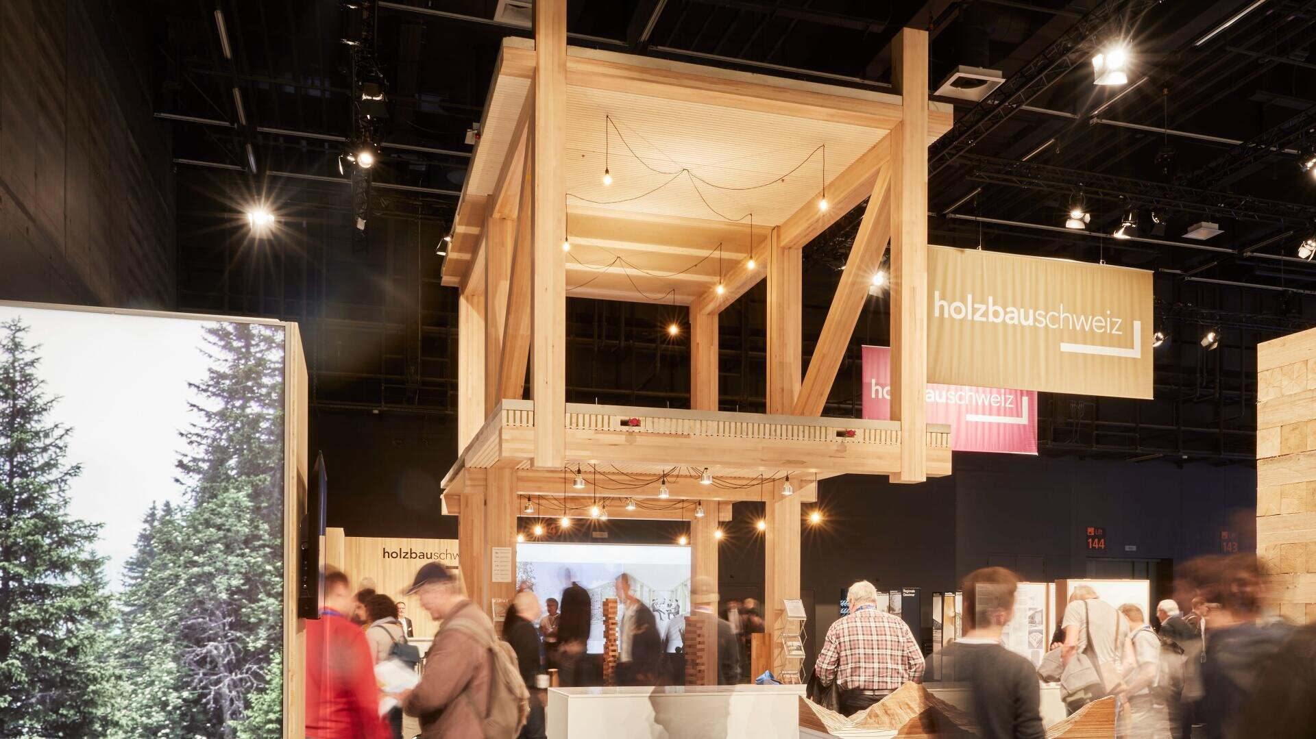 Eine belebte Indoor-Ausstellung mit einer großen Holzkonstruktion auf Stelzen, um die herum Menschen spazieren und sich versammeln; auf Bannern steht "holzbauschweiz". Auf der linken Seite ist ein Foto von Bäumen zu sehen.