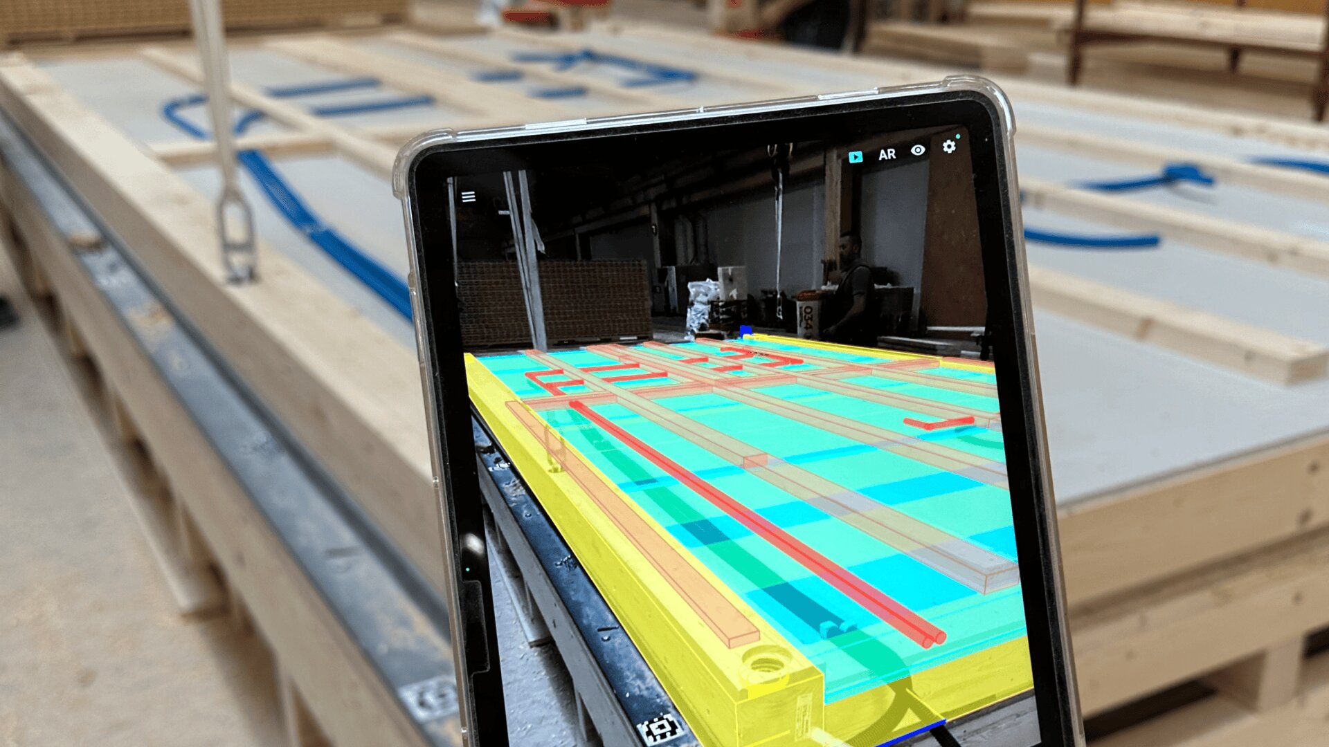 Une tablette affiche une superposition en réalité augmentée de formes et de lignes colorées sur un panneau de construction en bois, correspondant à sa disposition dans un atelier avec d'autres composants en bois assemblés en arrière-plan.