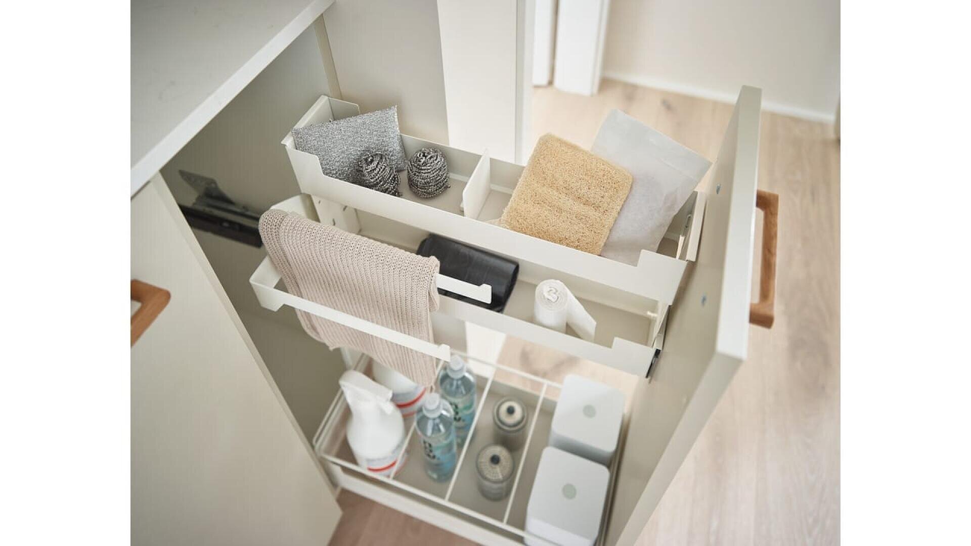 Une armoire de cuisine coulissante avec des étagères contenant des produits de nettoyage, des chiffons, des éponges, des flacons pulvérisateurs et des brosses en métal, le tout bien rangé. L'armoire est dotée d'une poignée en bois et repose sur un sol en bois clair.